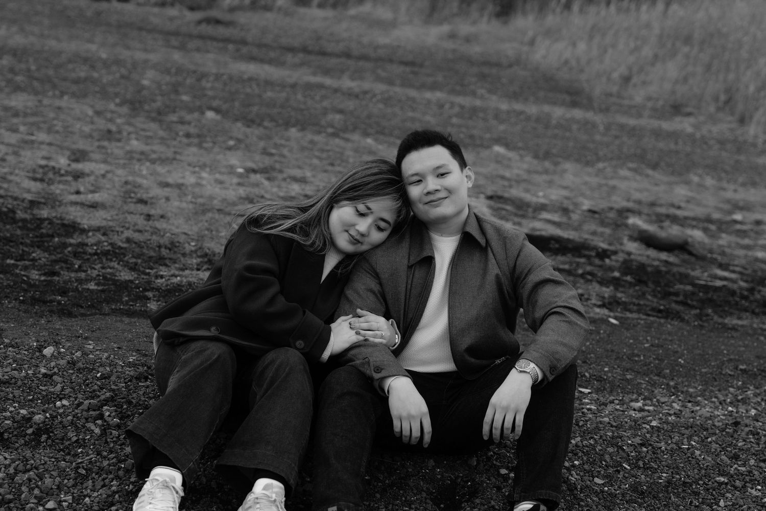Black and white photo of a couple sitting on rocky ground, woman leaning on mans shoulder, smiling outdoors.