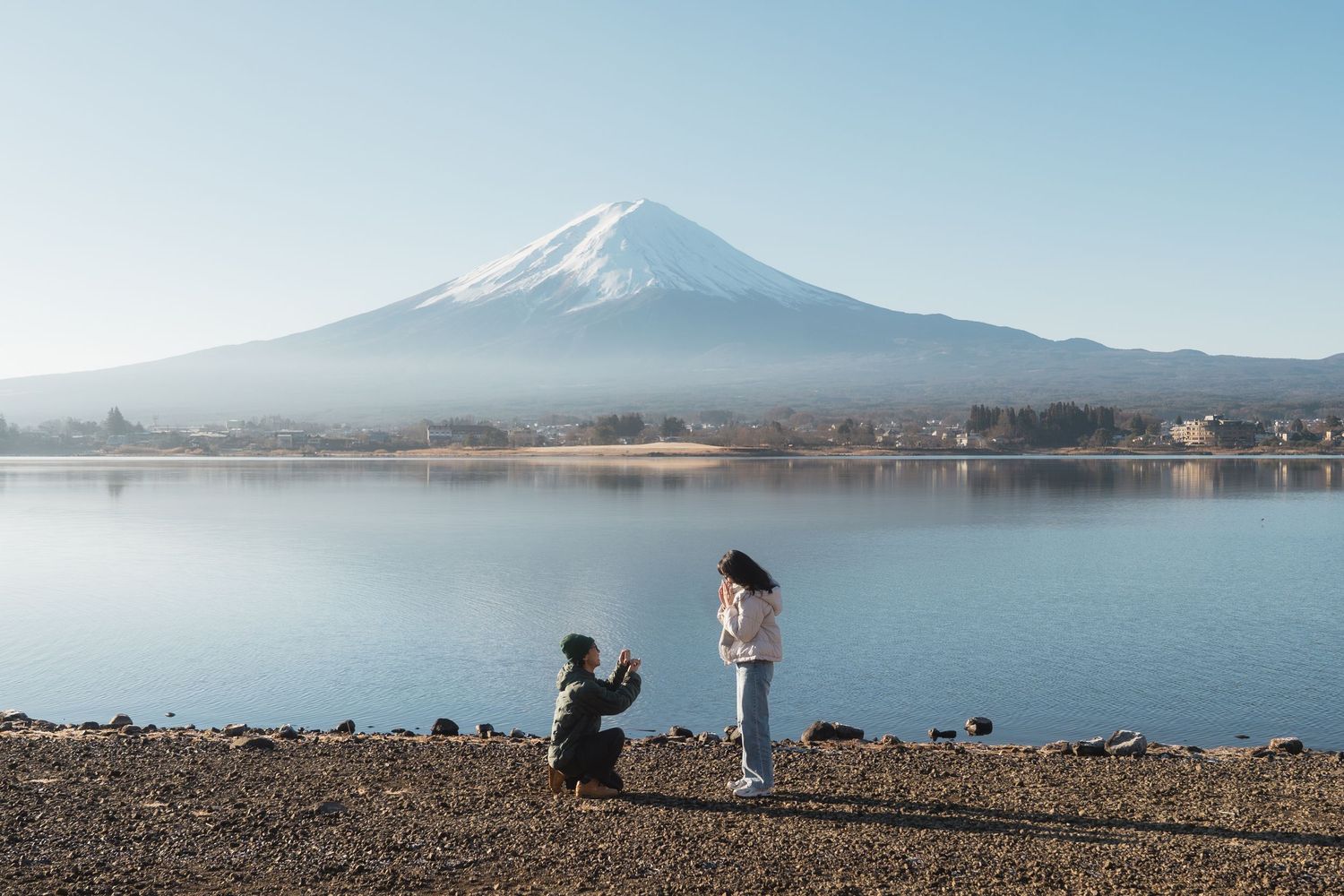 Man proposing on one knee to woman on rocky lakeshore with Mount Fuji in background.