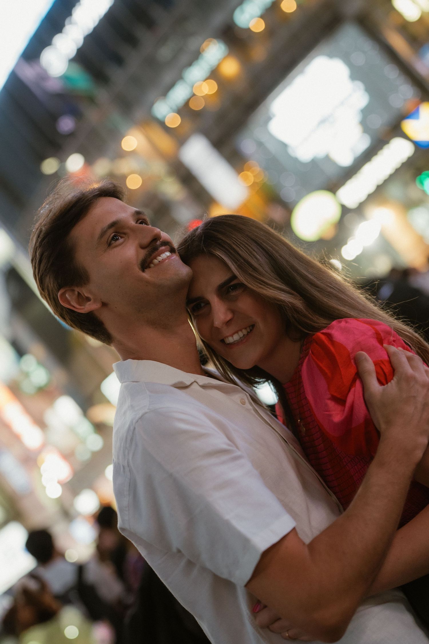 Happy couple embracing and smiling in a busy illuminated shopping arcade with bokeh lights.