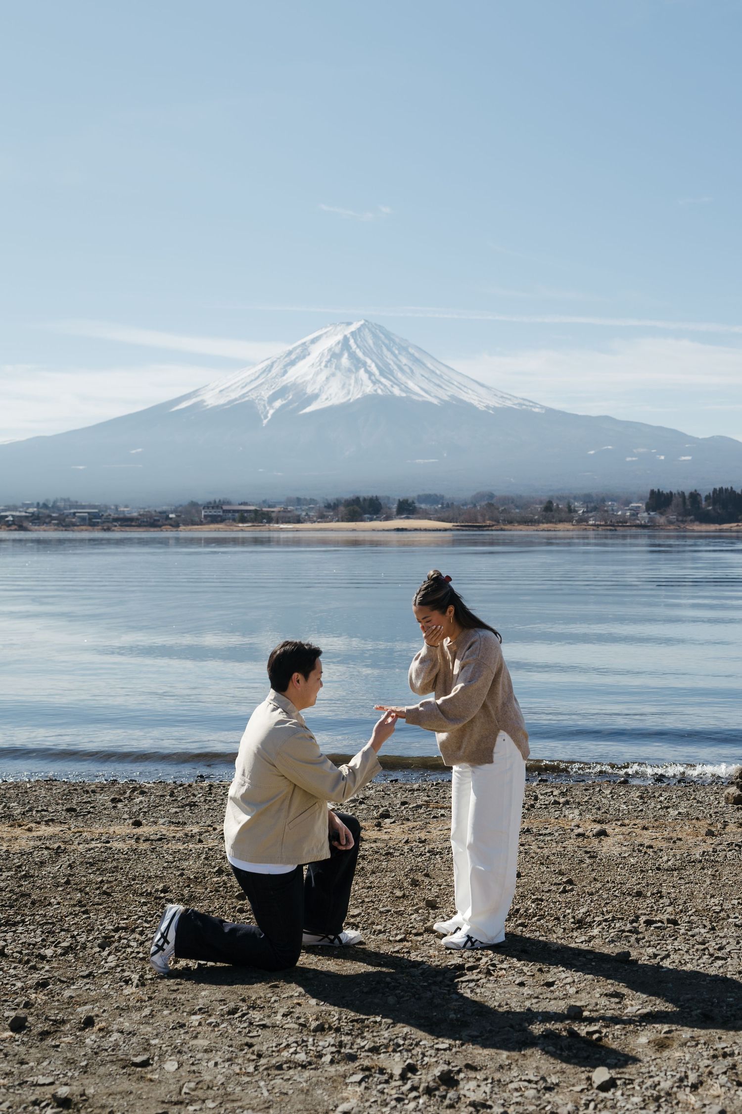 Man proposing on one knee to woman by Lake Kawaguchiko with Mount Fuji in background.