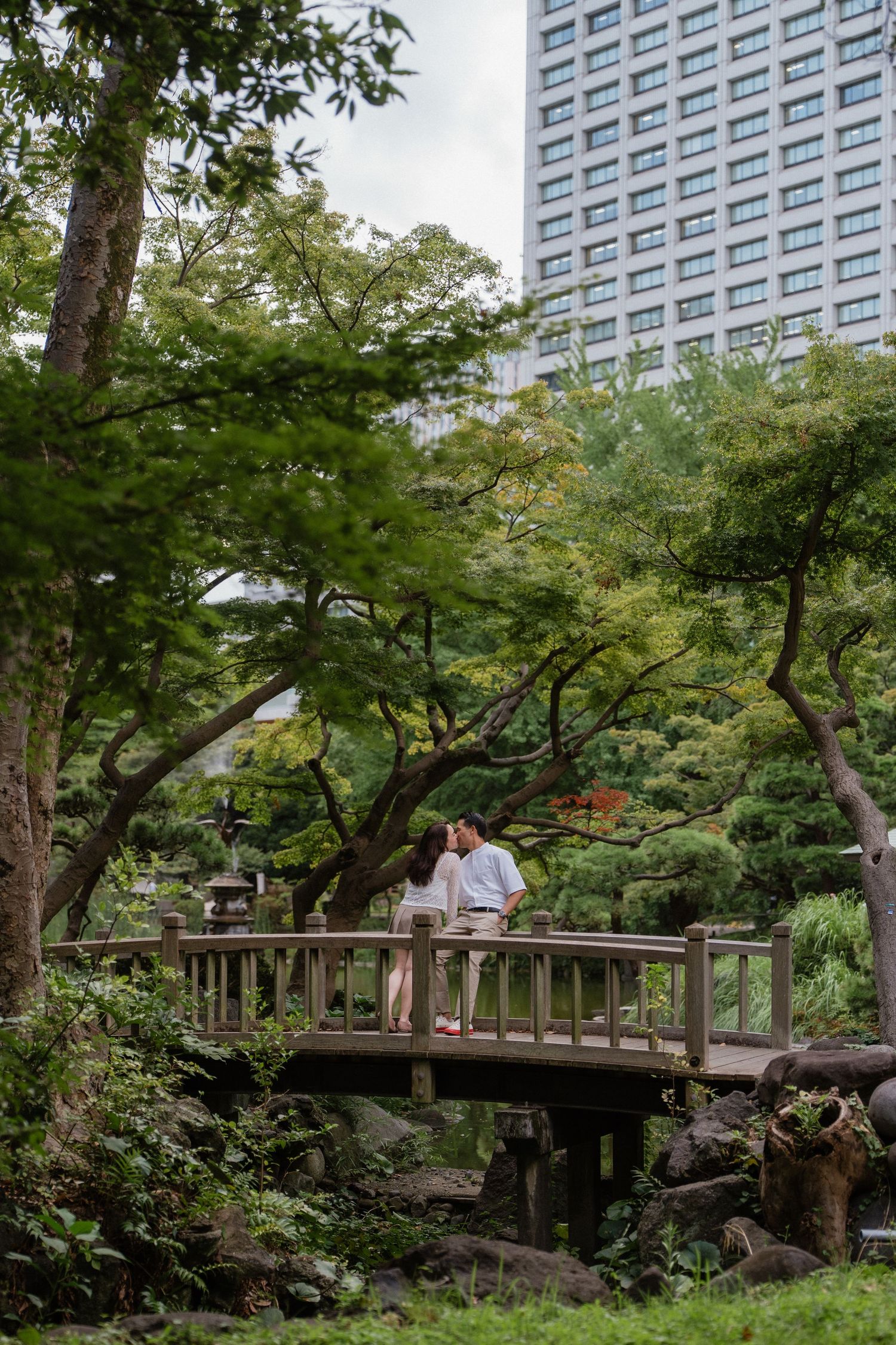 Couple embracing on a wooden bridge in a lush Japanese garden with a tall building in the background.
