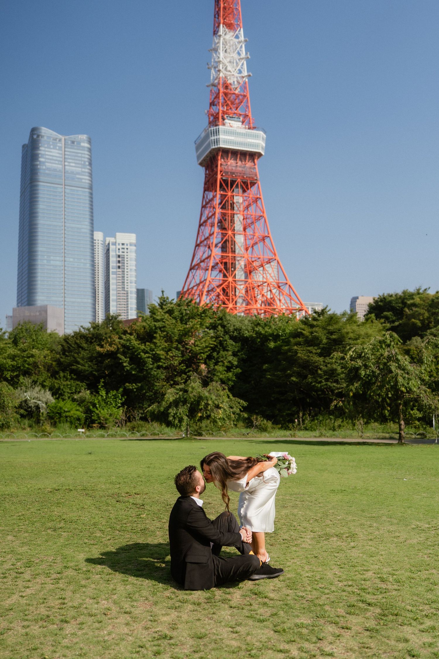 Couple sharing a romantic kiss on grass in front of Tokyo Tower on a clear sunny day.