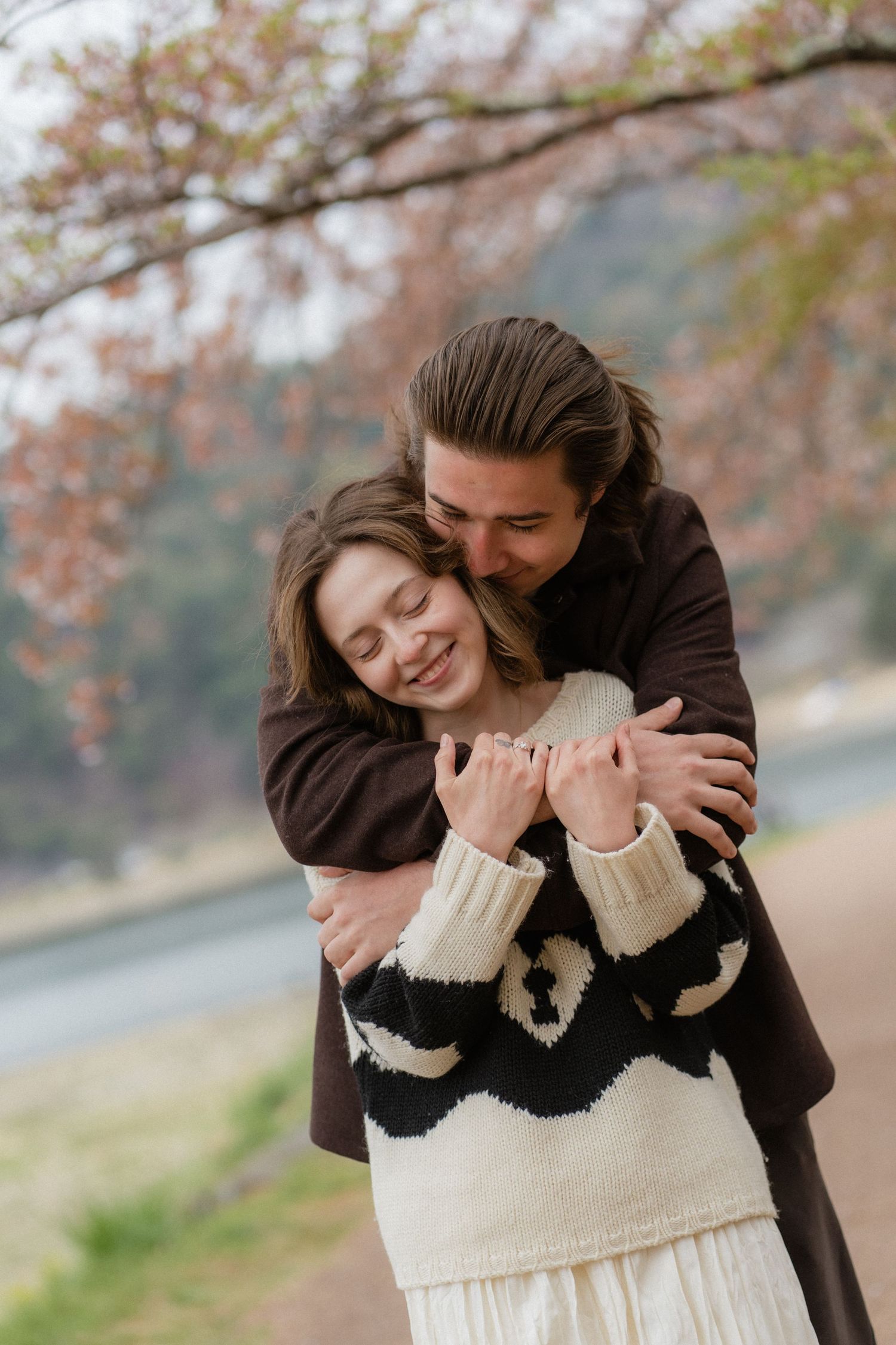 A couple embraces under cherry blossom trees, woman in patterned sweater smiling as man hugs her from behind.