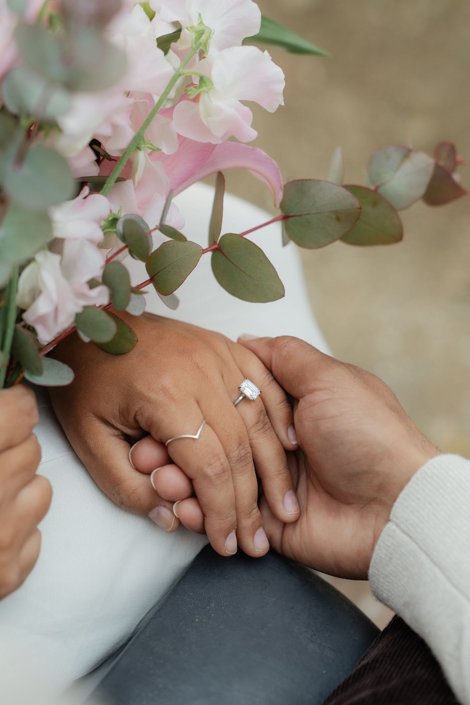 Close-up of couple holding hands with engagement ring and pink floral bouquet with eucalyptus leaves.