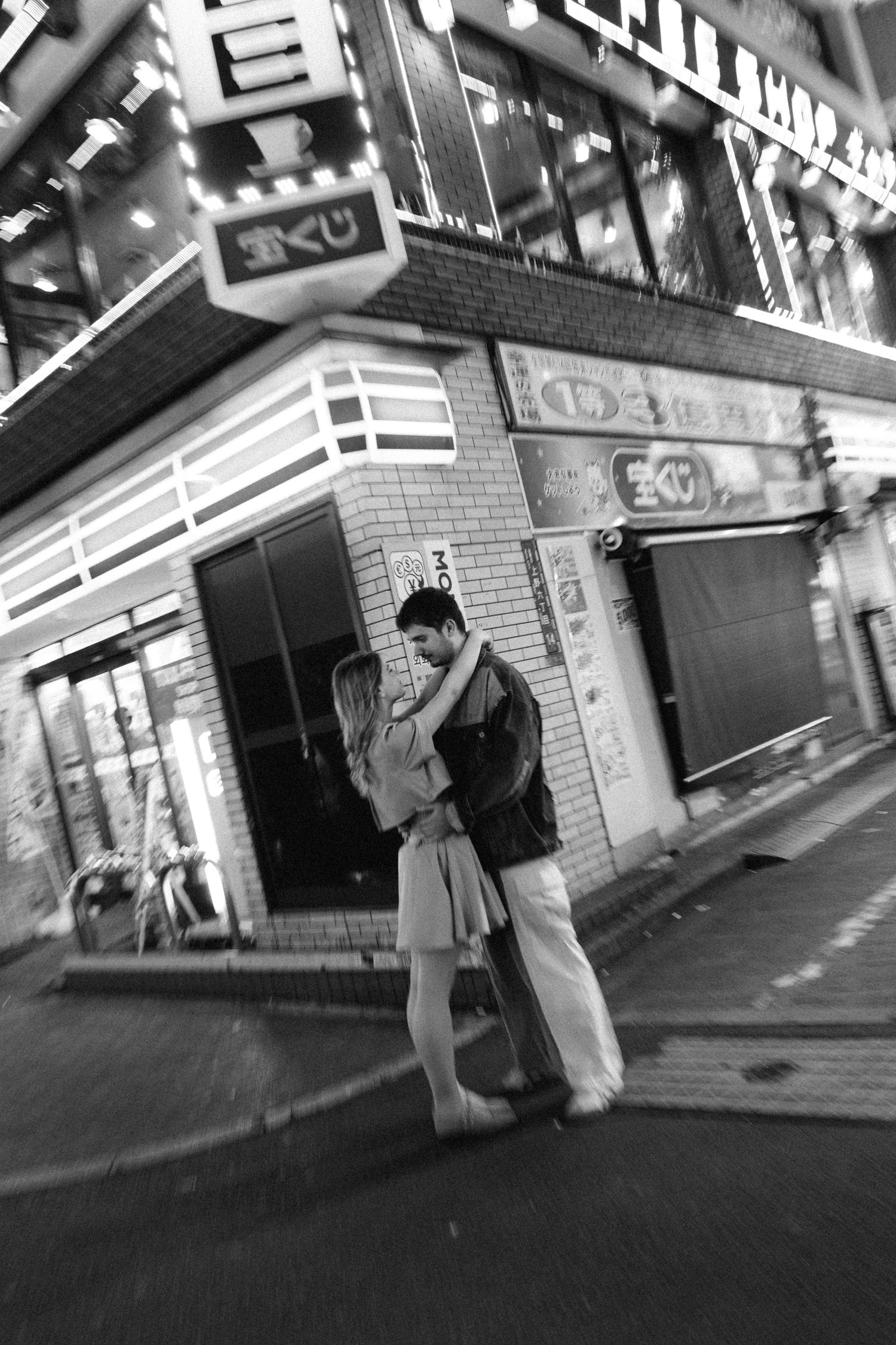 Black and white photo of a couple embracing on a street in Japan at night, with neon signs and storefronts behind them.
