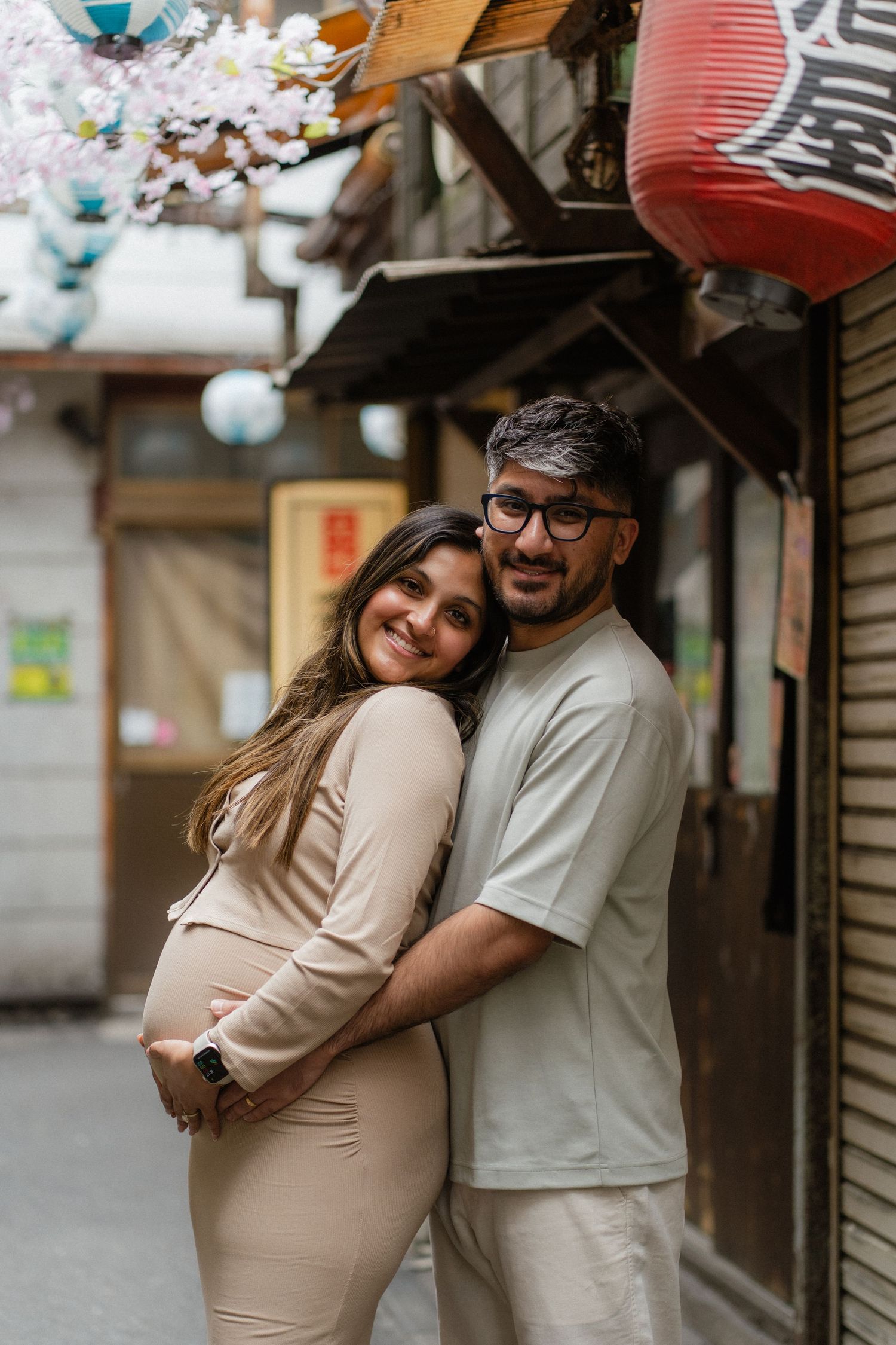 Happy pregnant couple embracing on a Japanese street with cherry blossoms and red lantern backdrop.
