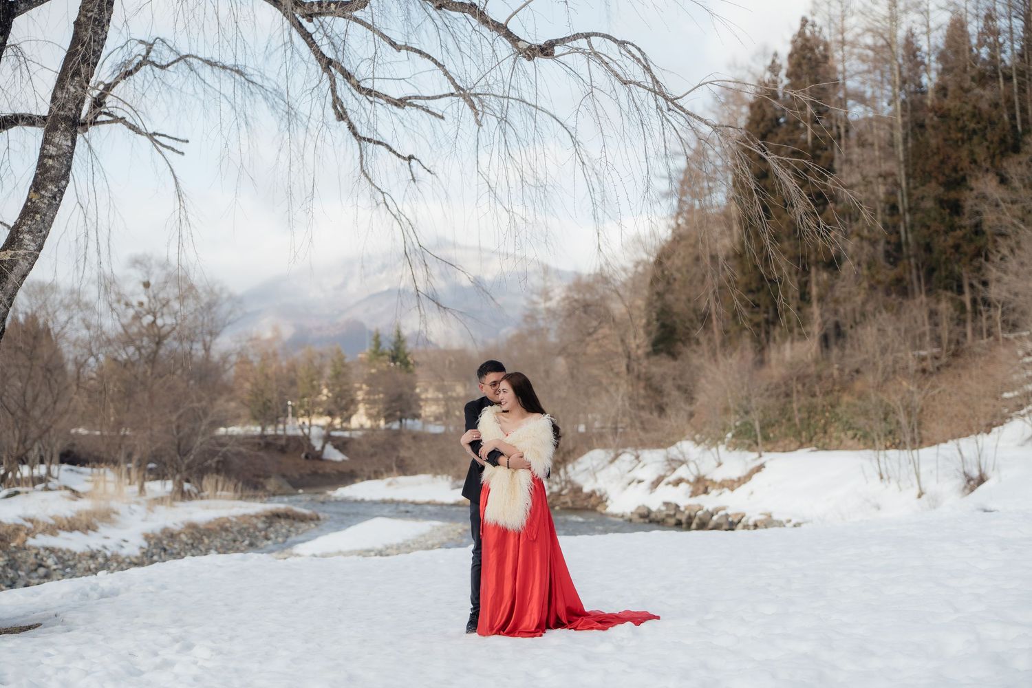 Couple embracing in snowy winter landscape wearing red gown and black suit with mountains in background.