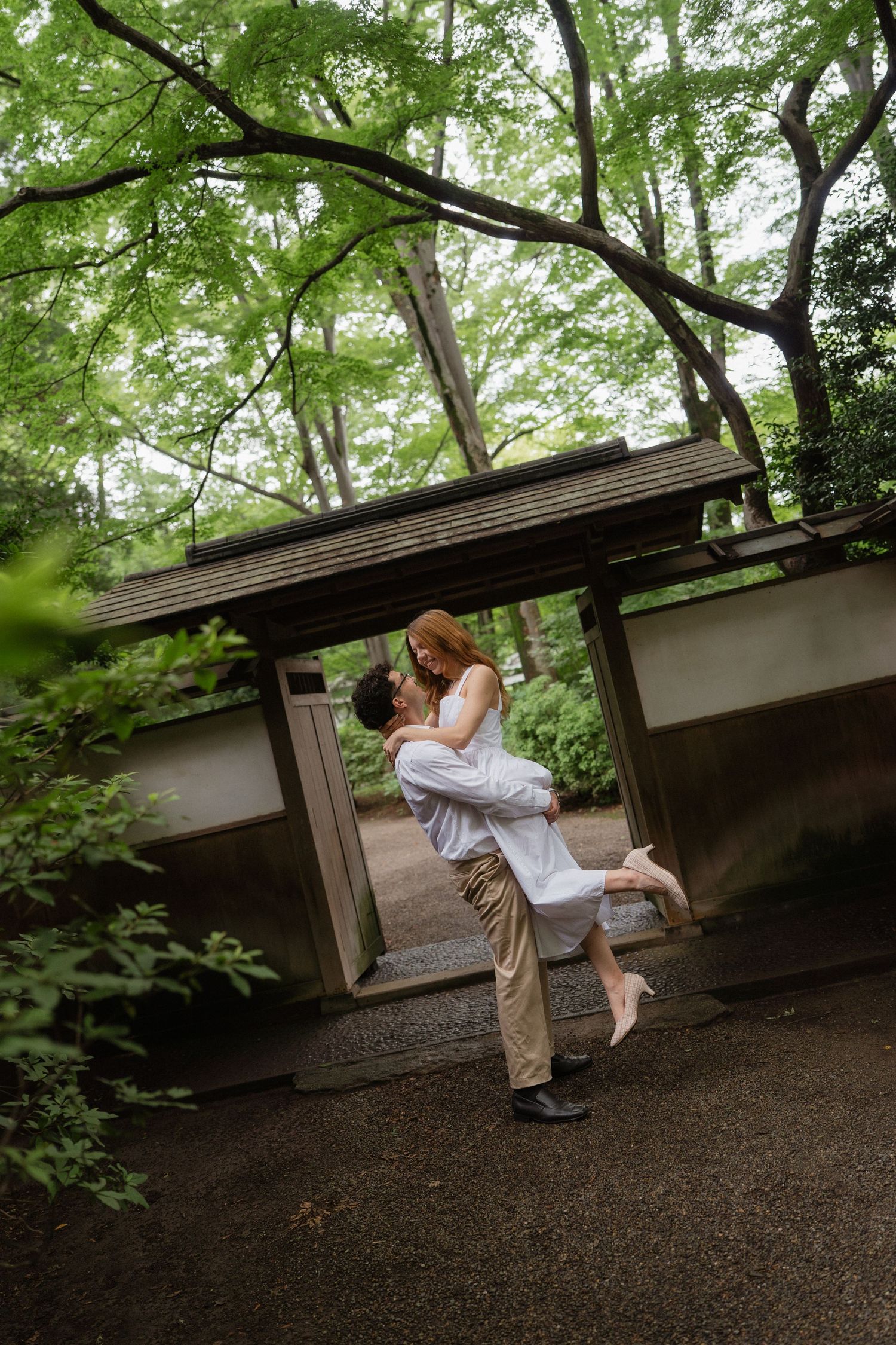 Couple embracing under a traditional Japanese garden gate surrounded by lush green trees.