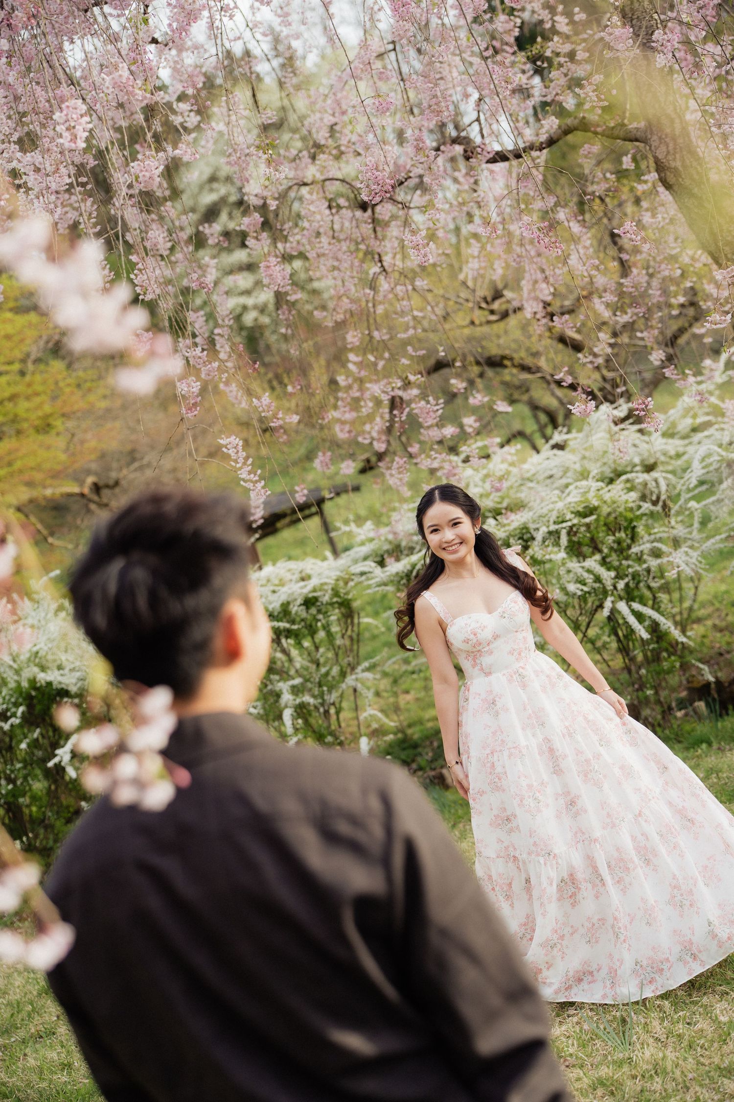 Couple in cherry blossom garden, woman in floral ball gown smiling at man in black shirt during spring photoshoot.