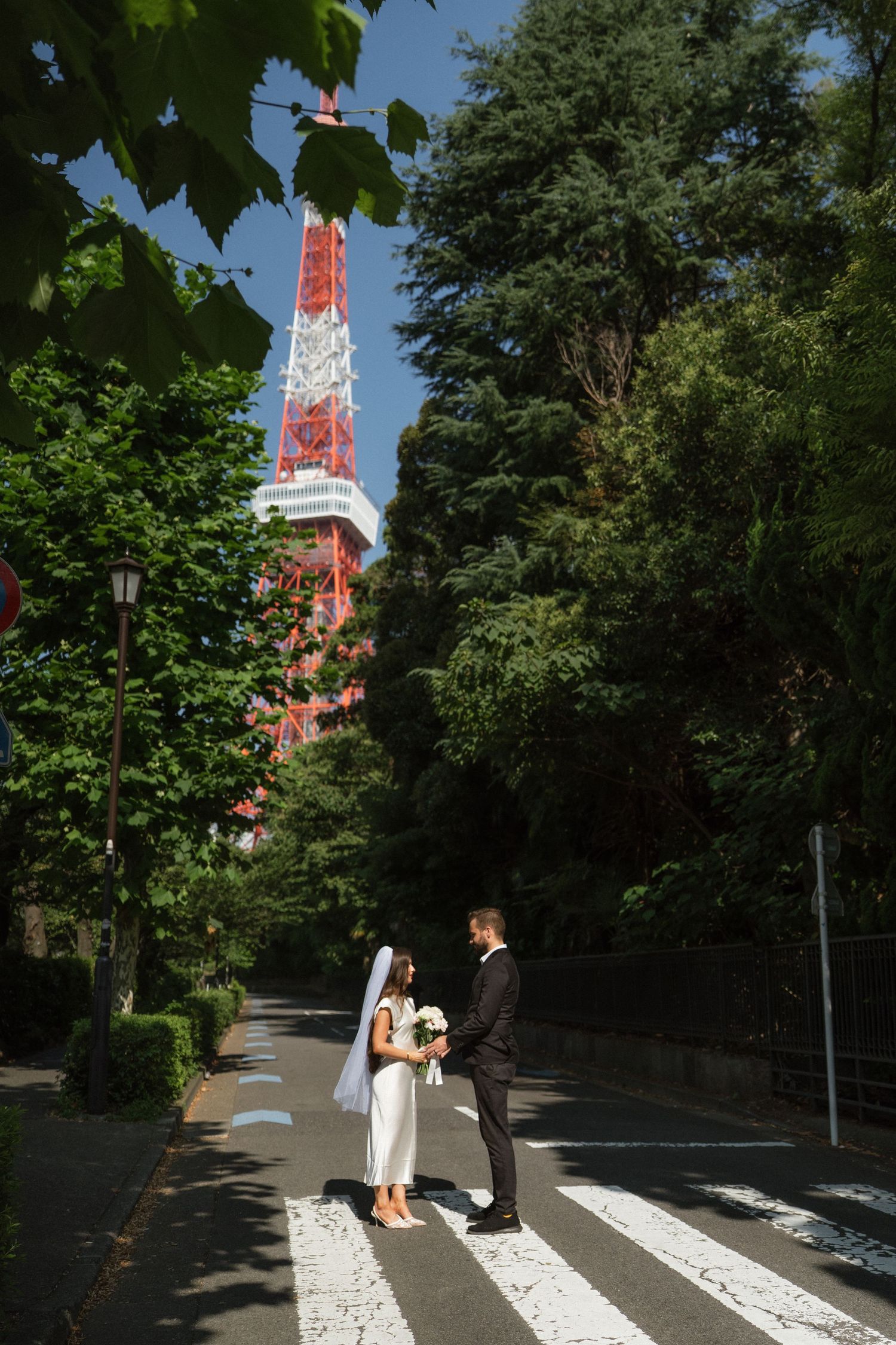 Bride and groom stand on crosswalk near Tokyo Tower surrounded by lush green trees on a sunny day.