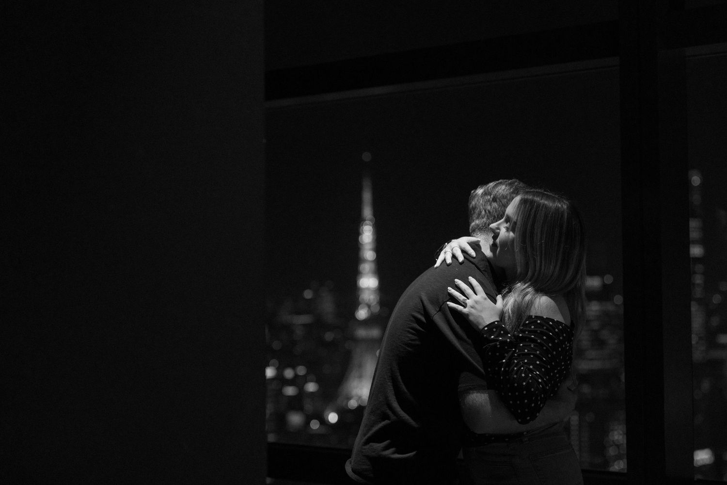 Black and white photo of a couple embracing by a window with the Tokyo Tower illuminated in the night skyline.