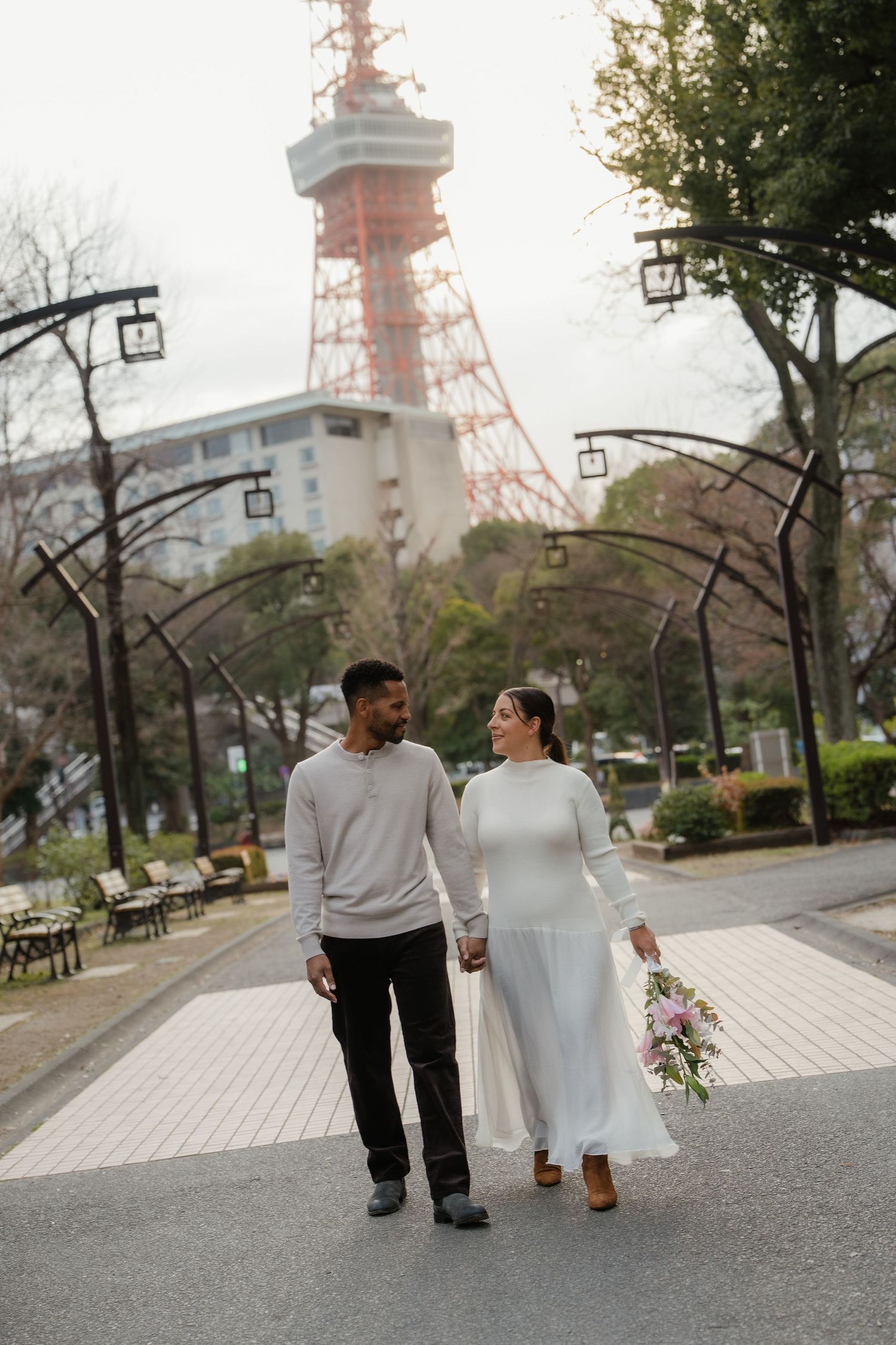 Couple holding hands in front of Tokyo Tower, woman in white dress holding bouquet, man in gray sweater.
