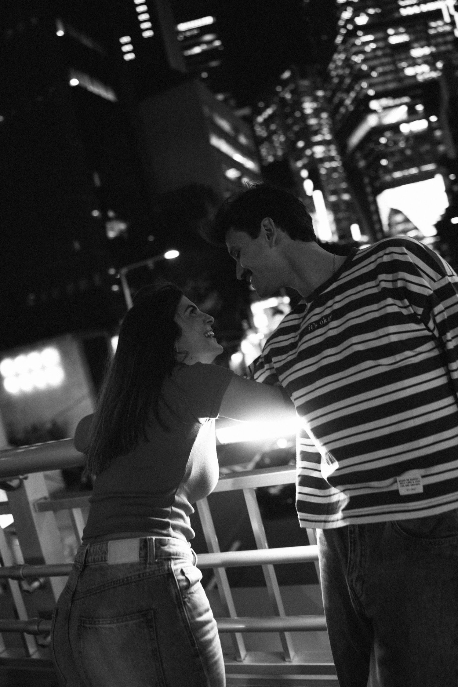 Black and white photo of a romantic couple gazing at each other at night with city lights in the background.