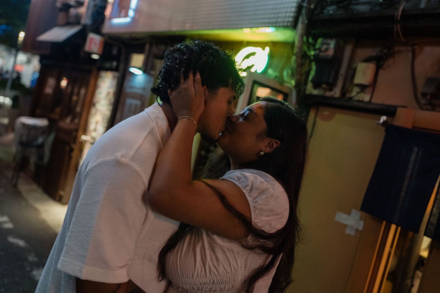 Couple sharing a romantic kiss on a vibrant night street, surrounded by glowing signs and urban ambiance.