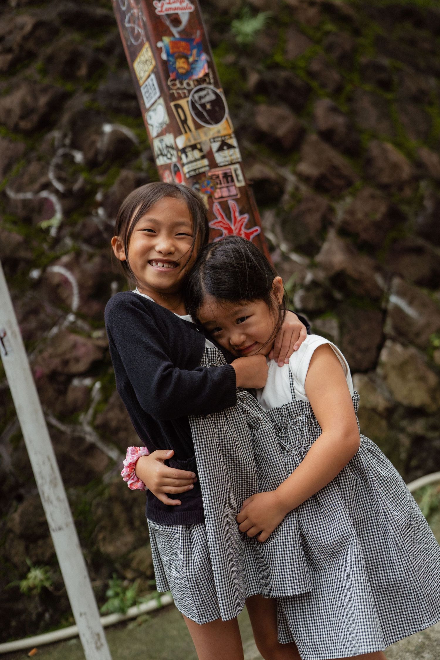 Two young Asian girls hugging and smiling outdoors near a rocky wall with stickers.