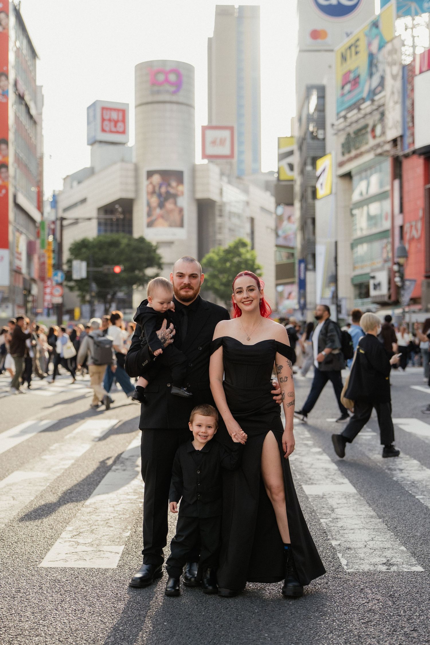 Family in matching black outfits posing at Shibuya Crossing in Tokyo, with city buildings and crowds in the background.