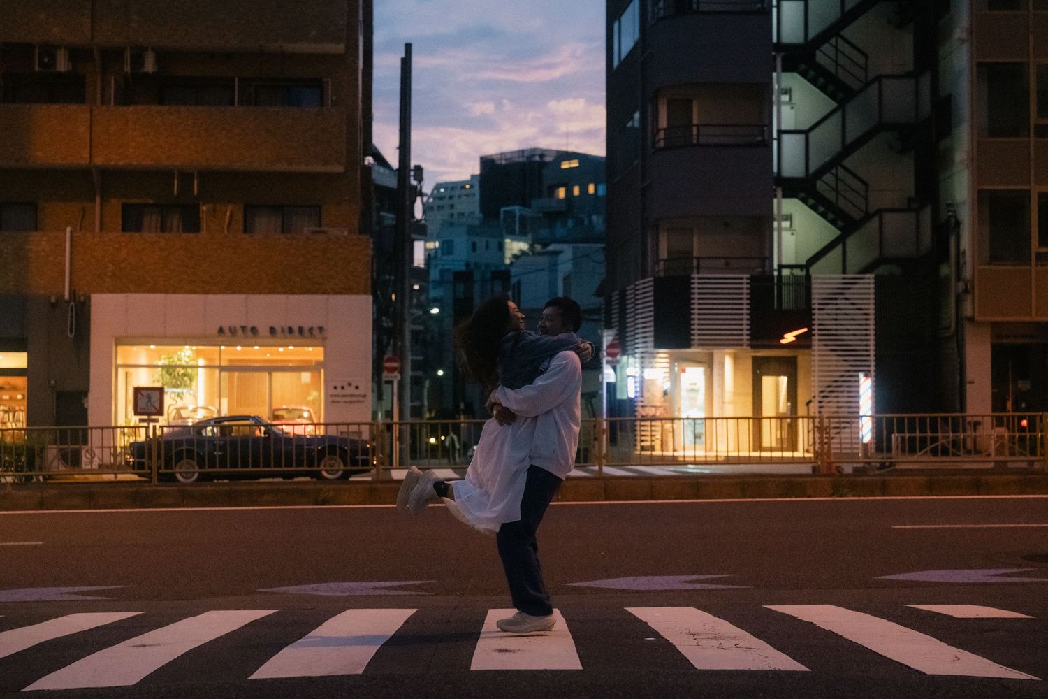 Couple embracing on a crosswalk at dusk in a Tokyo city street with illuminated buildings.