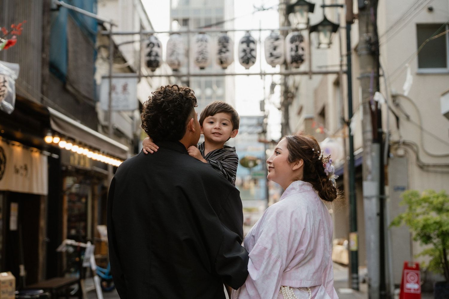 A family with a toddler walks through a narrow Japanese alley, the woman wearing a white kimono.
