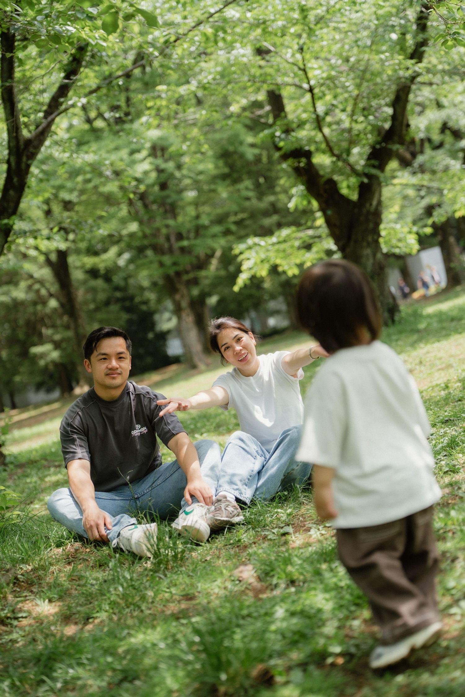 Asian family relaxing in green park, parents sitting on grass while young child runs toward them.