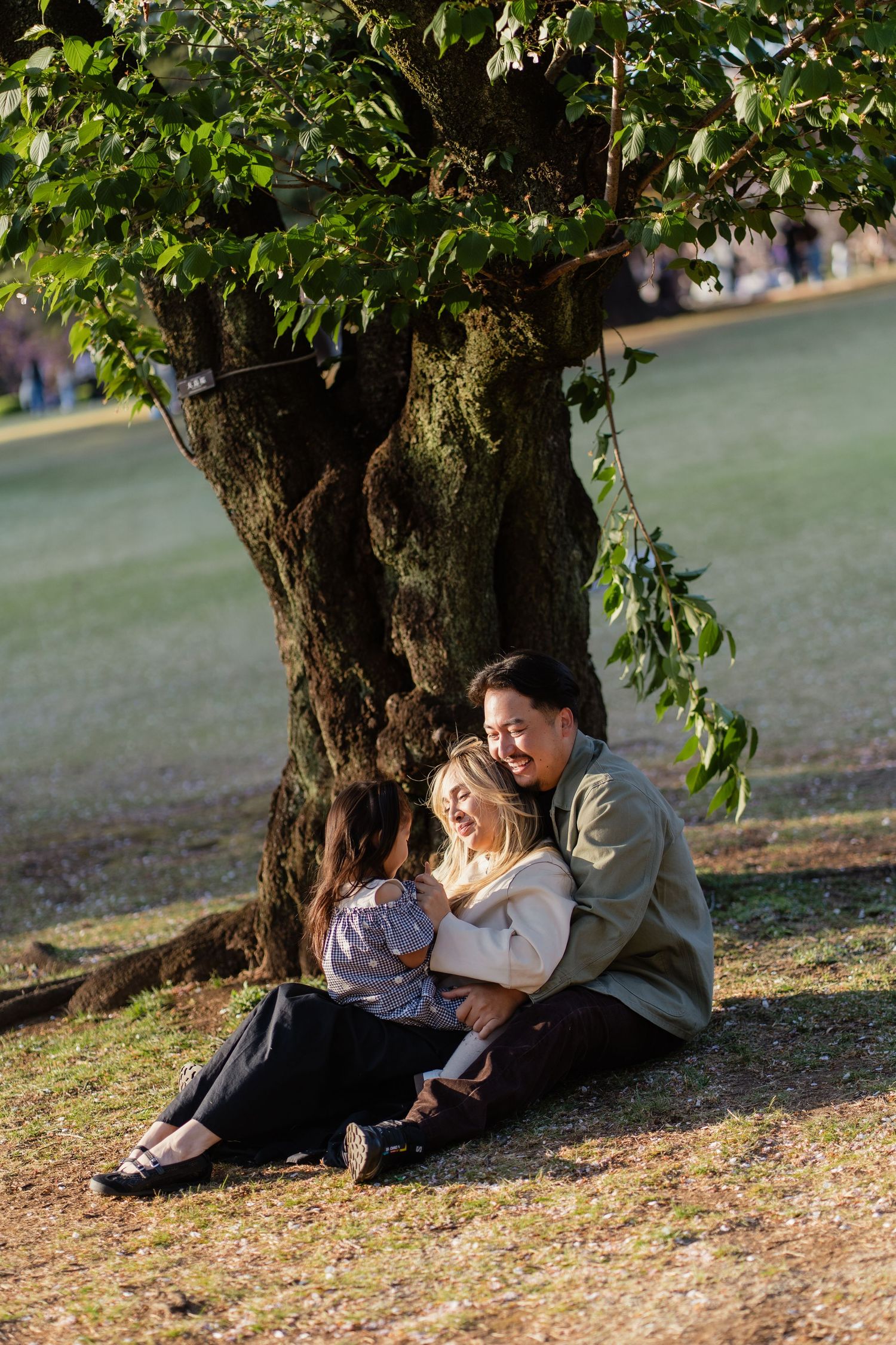 Family of three sitting together under a large tree in a sunny park, sharing a joyful moment.