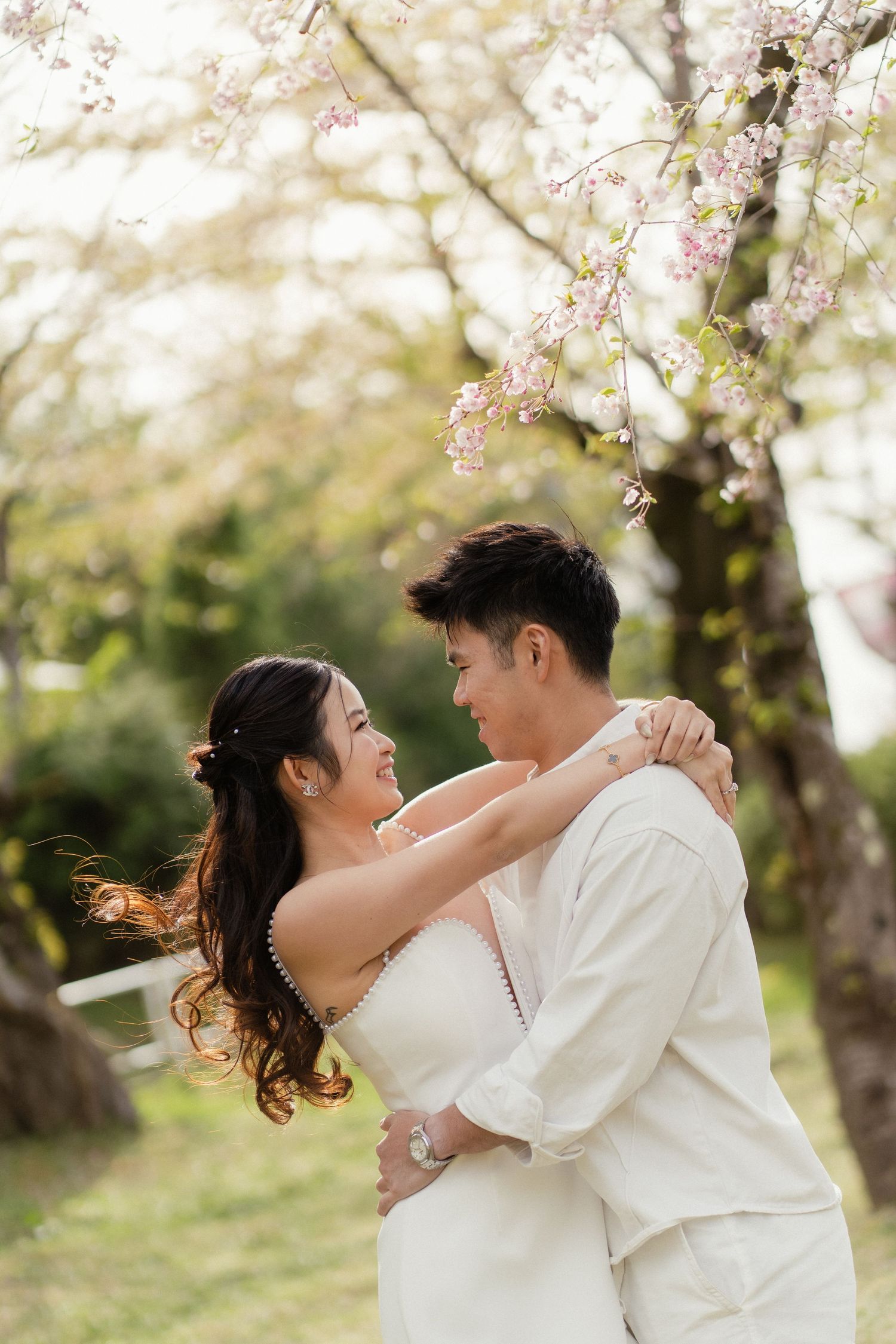 A couple in white wedding attire embraces under blooming cherry blossom trees, smiling at each other in a romantic garden.
