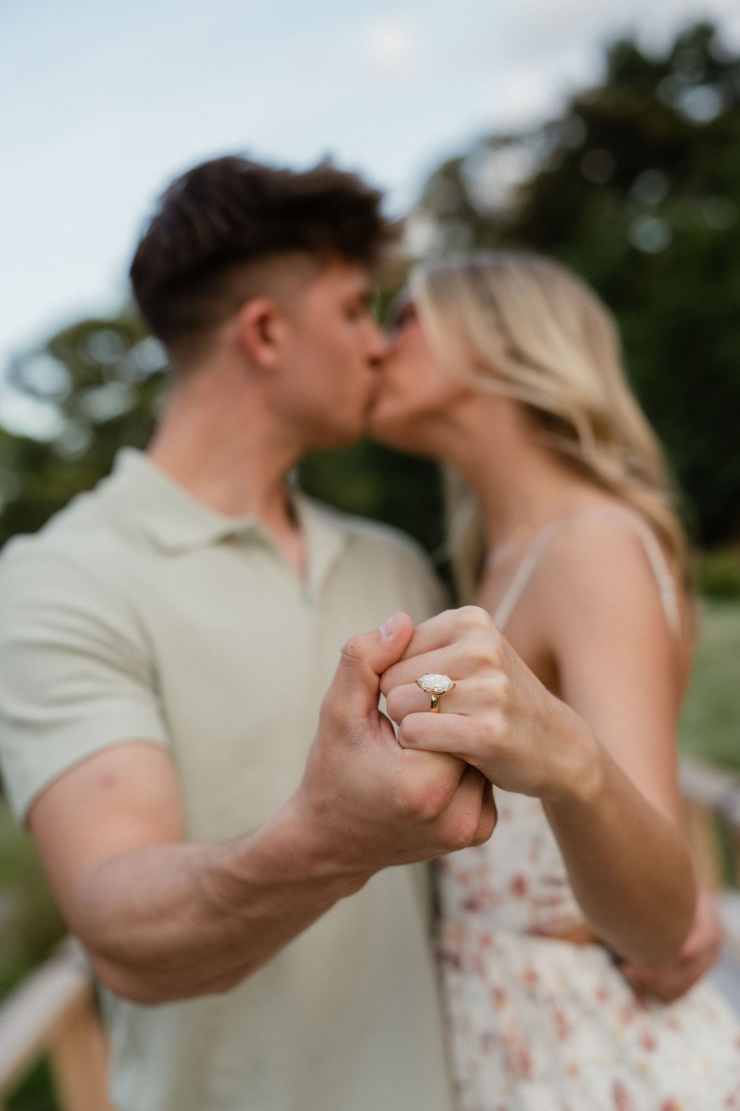 Engaged couple kissing outdoors while displaying a diamond engagement ring on her finger.