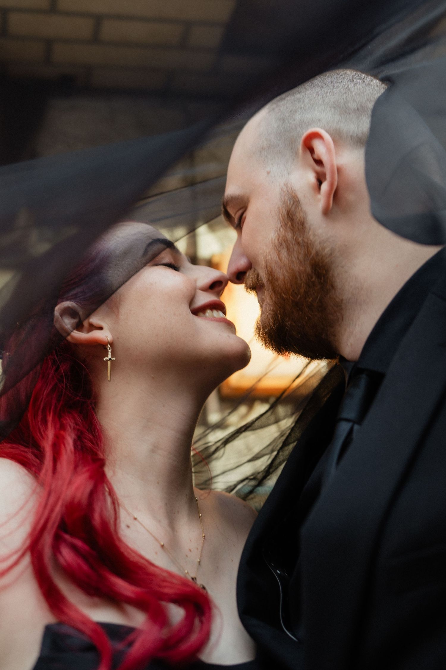 A couple shares a romantic moment under a black veil, the bride with red hair smiling as the groom leans in for a kiss.