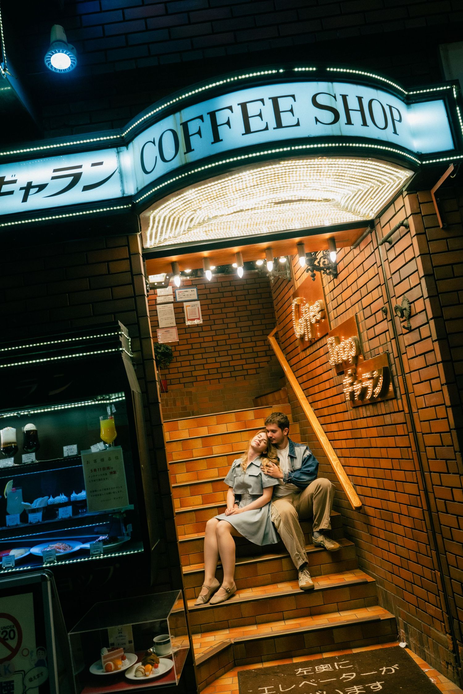 Couple sitting on steps outside a neon-lit Japanese coffee shop at night, surrounded by brick walls.
