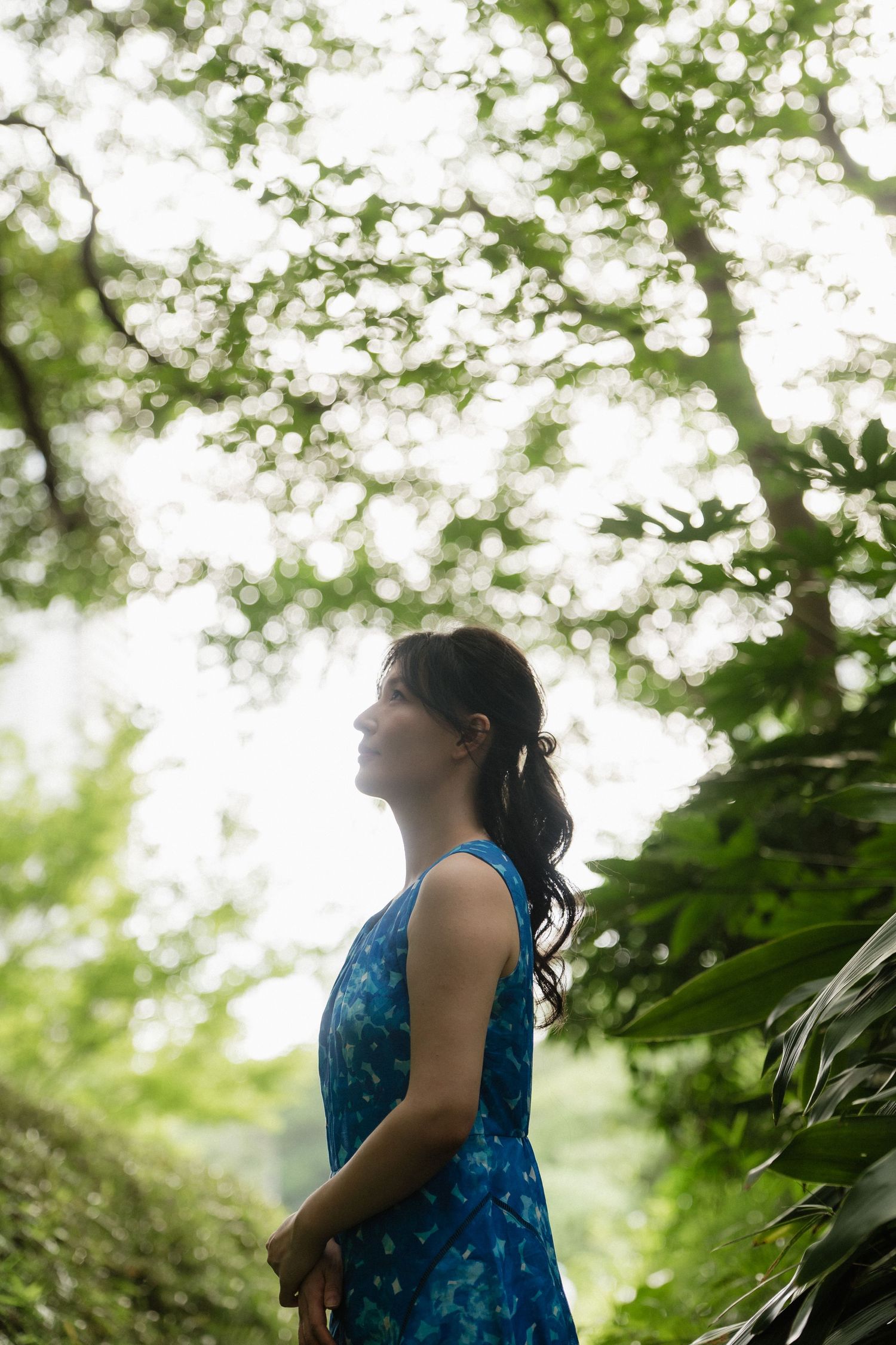A young woman in a blue floral dress stands peacefully among lush green trees in a serene forest setting.