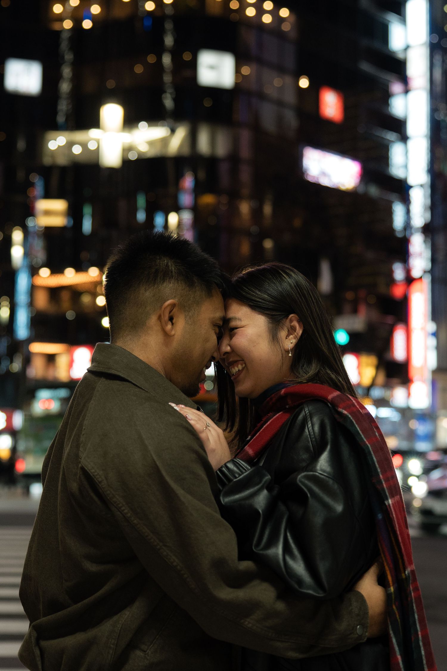 Couple sharing a romantic moment at night in a vibrant city, smiling with foreheads touching.