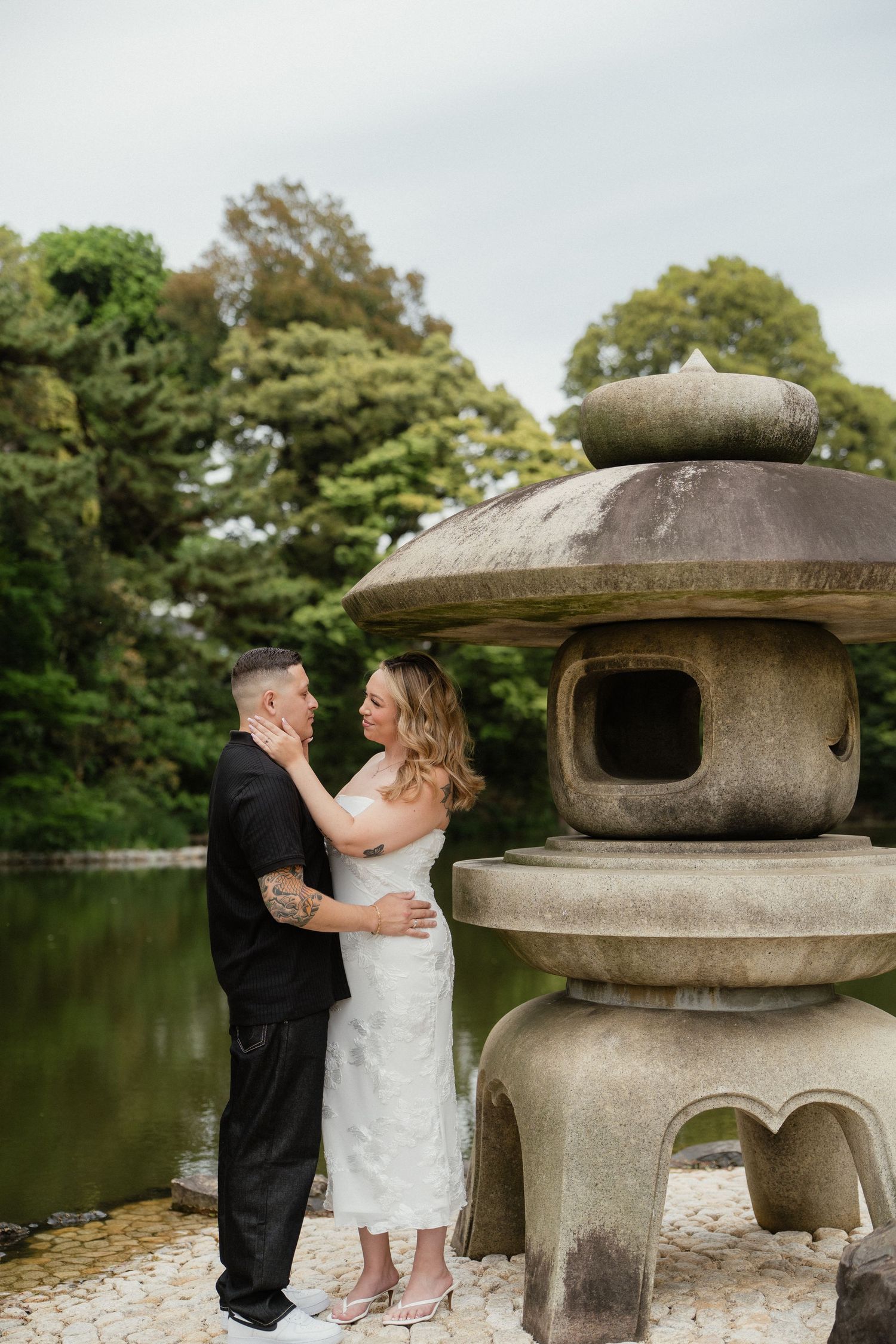 Couple embracing near a stone Japanese lantern by a serene pond, surrounded by lush green trees.