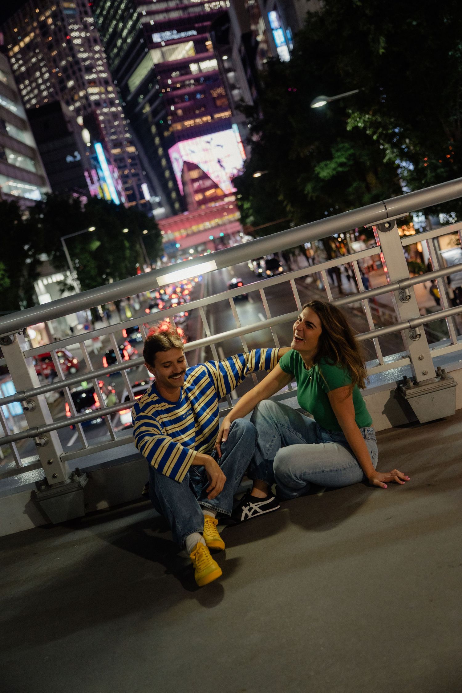 Couple sitting on urban bridge at night with illuminated city skyscrapers behind them, wearing casual colorful outfits.