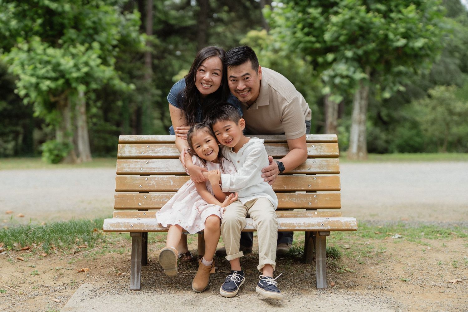 Happy Asian family of four posing together on a wooden park bench surrounded by lush green trees.