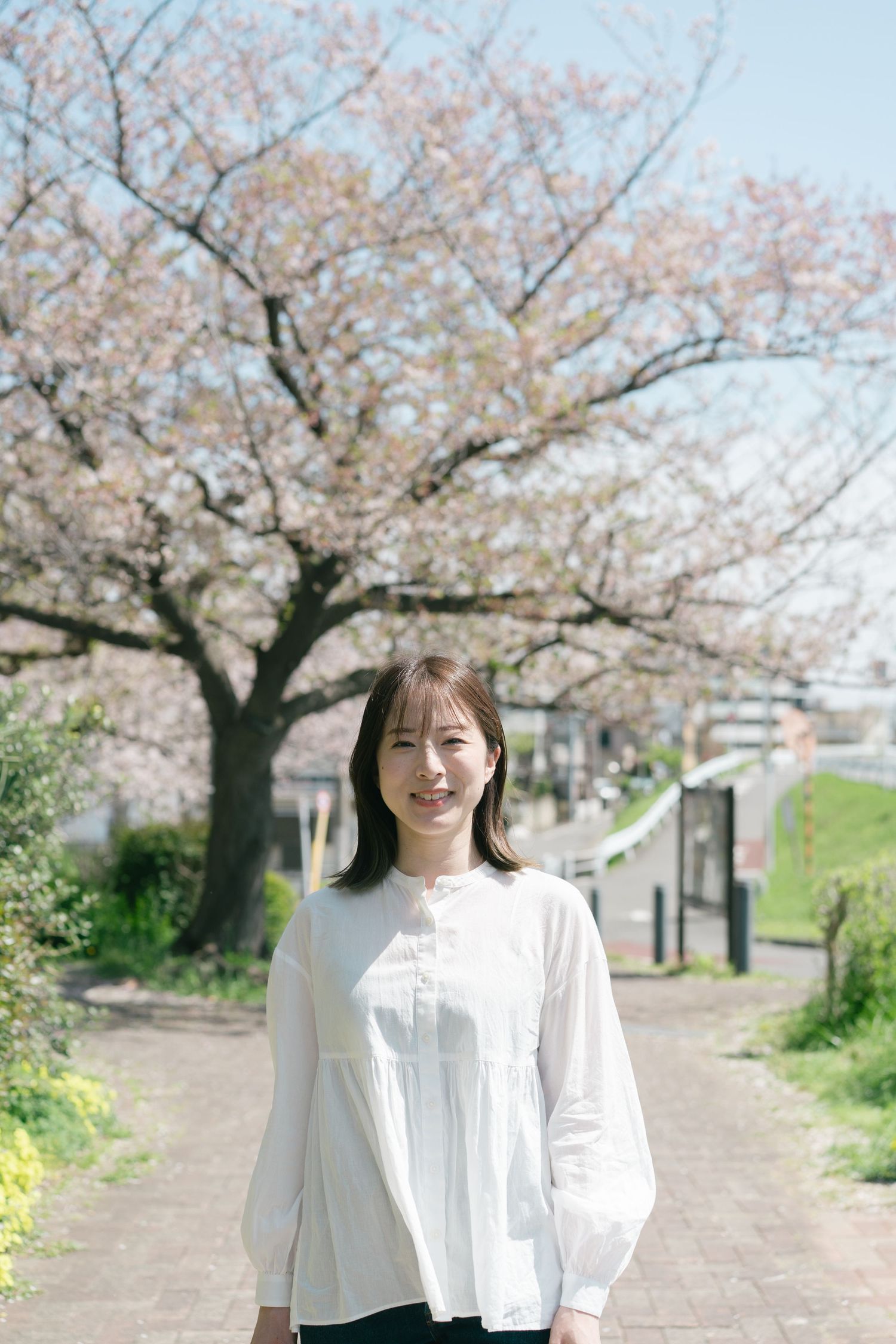 Smiling young Asian woman in white top standing on a path beneath blooming cherry blossom trees.