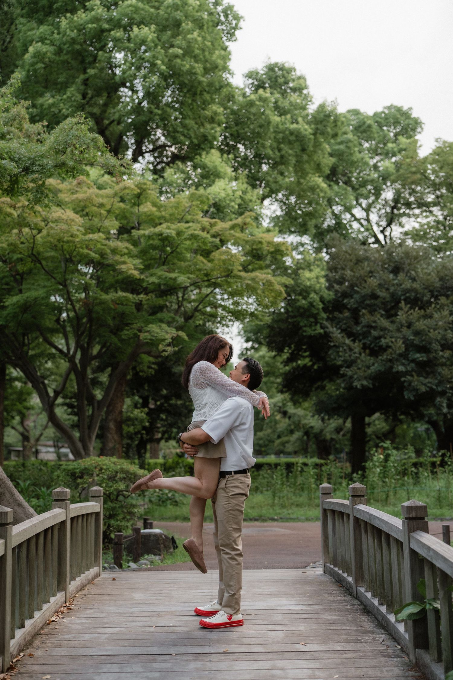 A couple shares a romantic moment on a wooden bridge, the man lifting the woman surrounded by lush green trees.