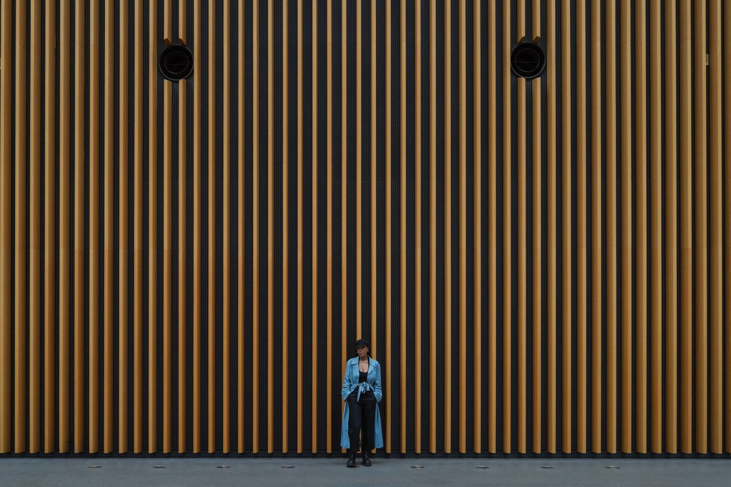 Person in blue jacket standing against a tall wooden slat wall with two dark vents above.