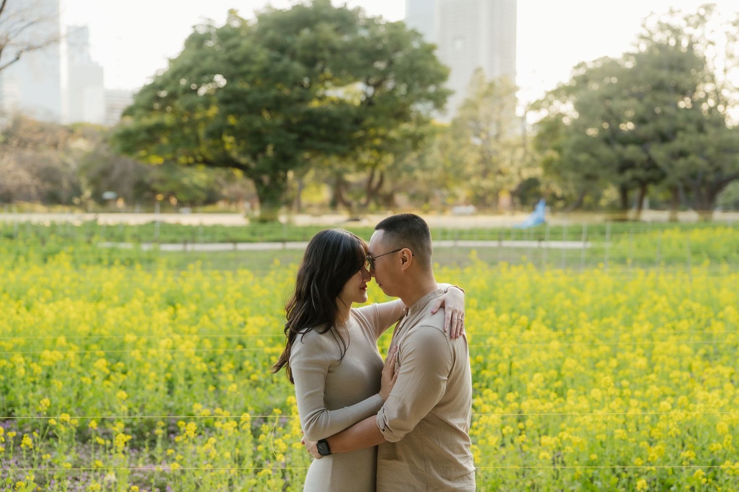 Couple embracing in a vibrant yellow flower field with lush green trees and city skyline in the background.
