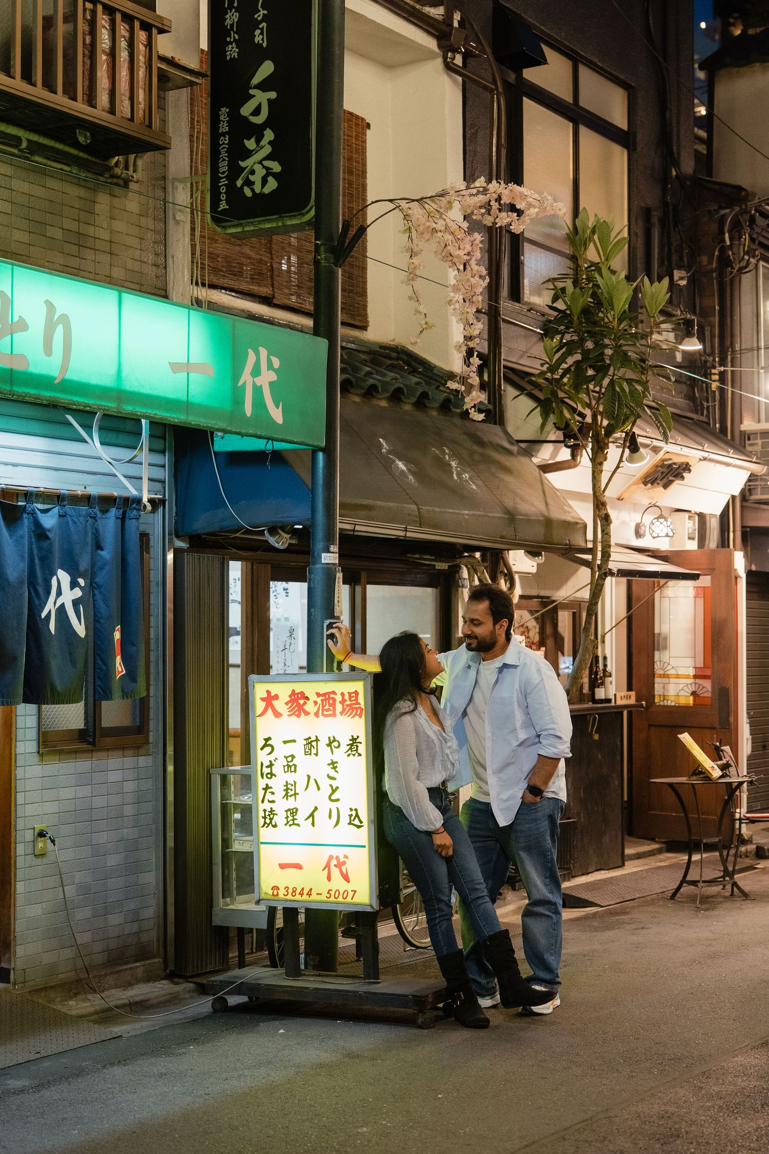 Couple smiling and holding hands on a Japanese street at night, surrounded by glowing restaurant signs.