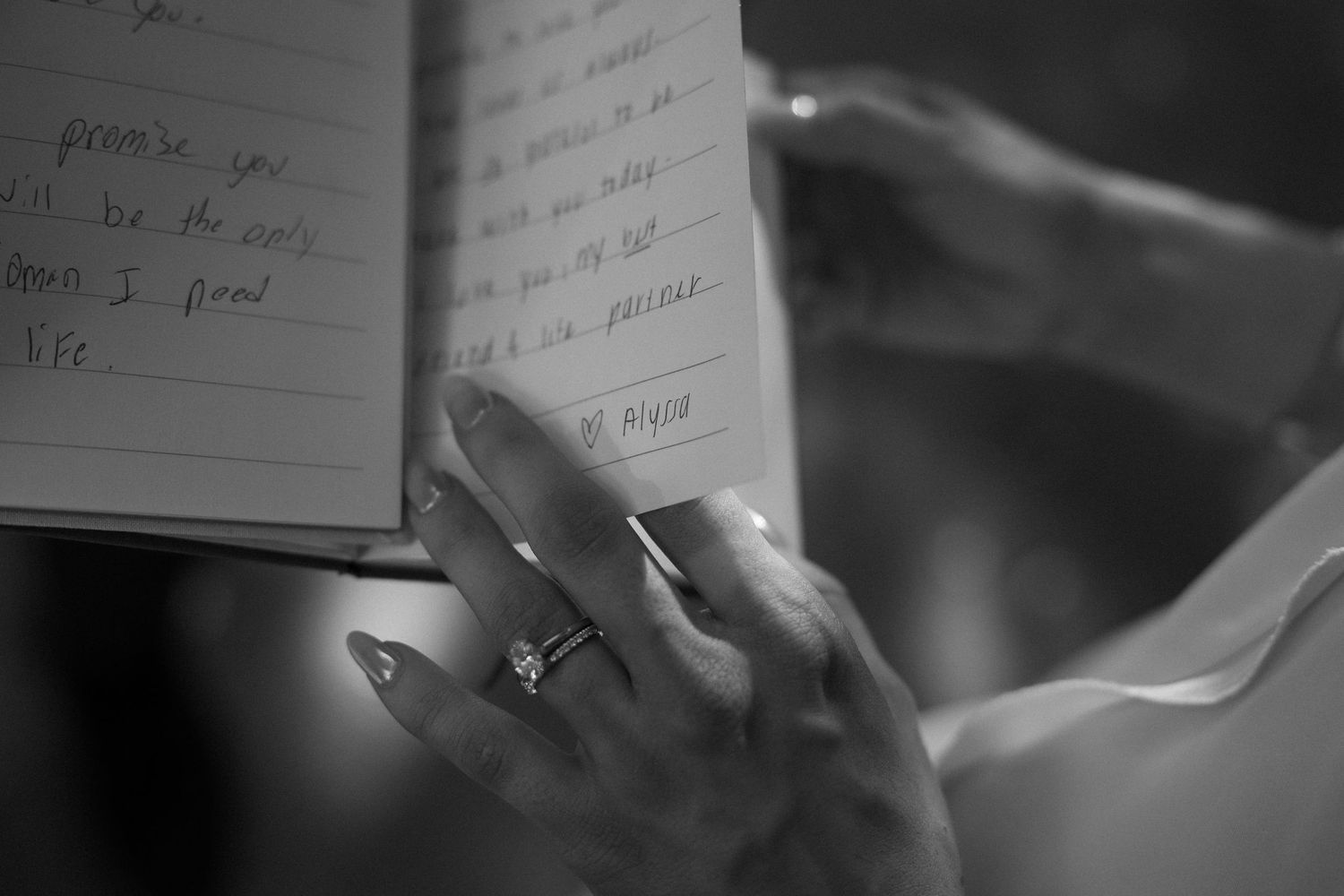 Close-up black and white photo of hands with engagement ring writing in a notebook with a pen.
