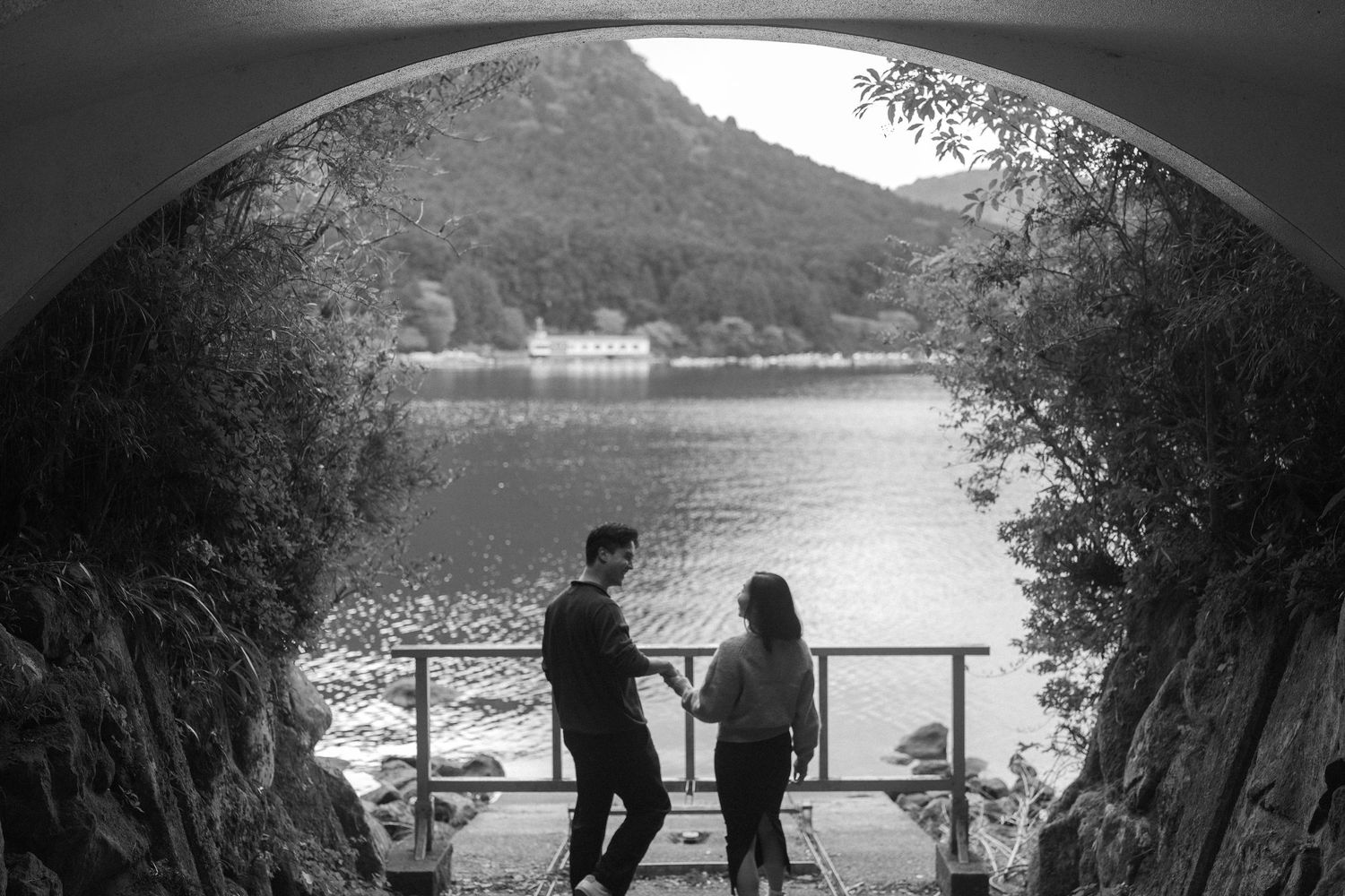 Black and white photo of a couple standing under an arch tunnel overlooking a serene lake and mountain landscape.