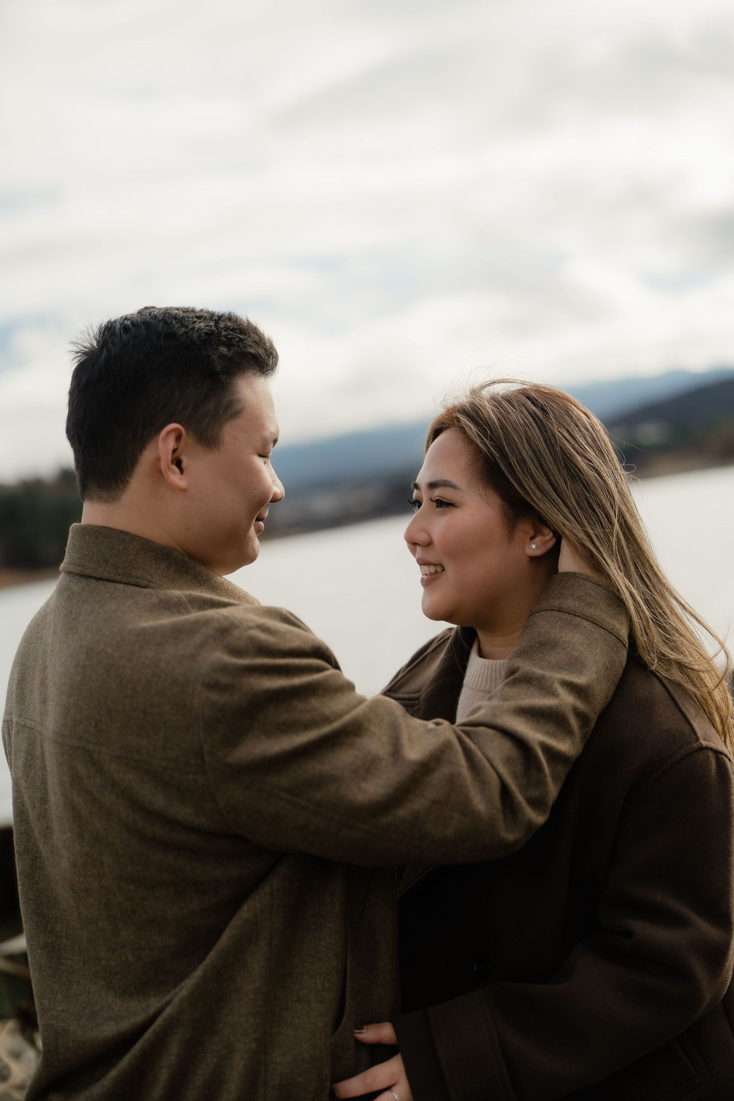 A couple embraces and smiles at each other near a lake on a cloudy day, both wearing brown coats.
