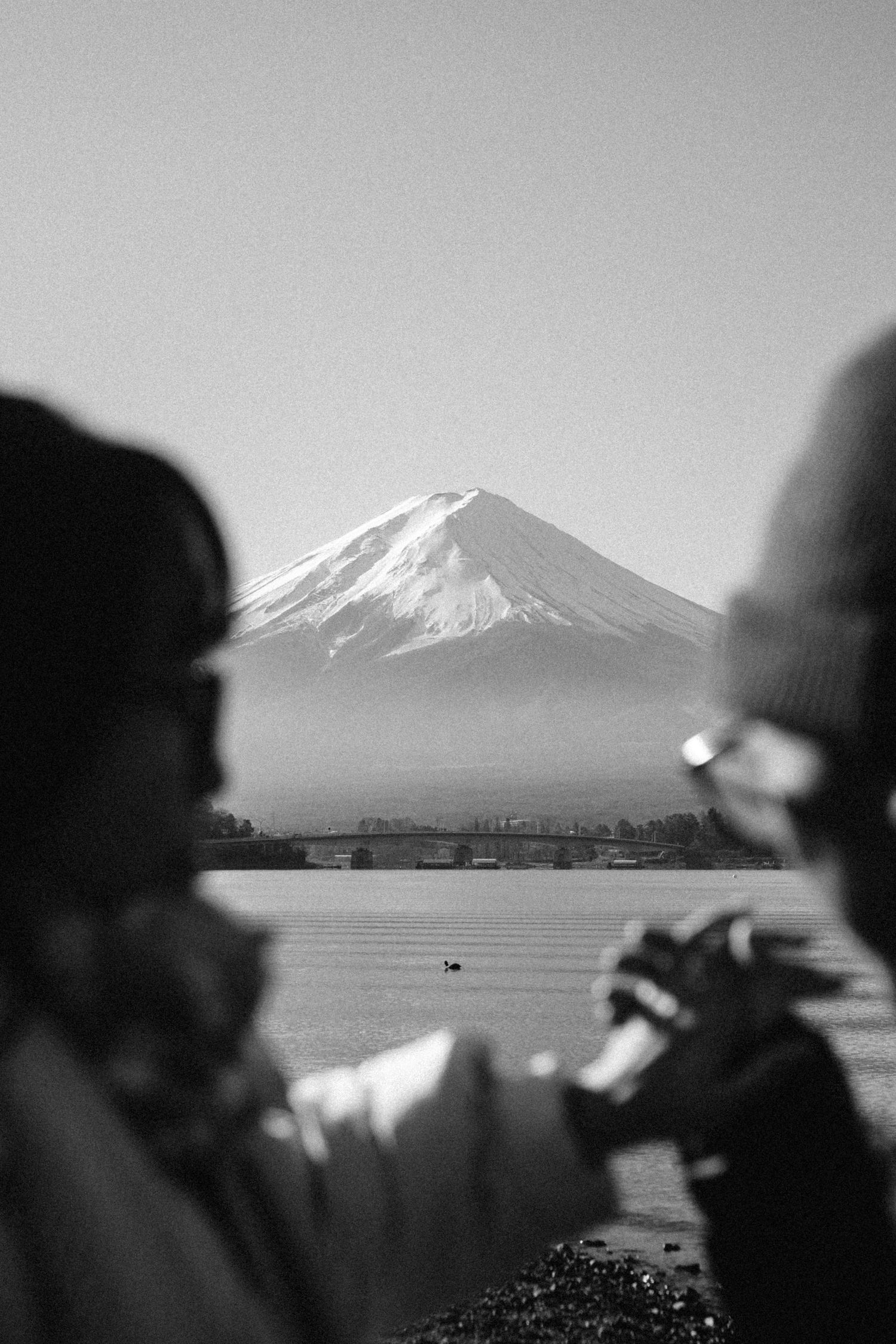 Black and white photo of two people photographing snow-capped Mount Fuji across a calm lake in Japan.
