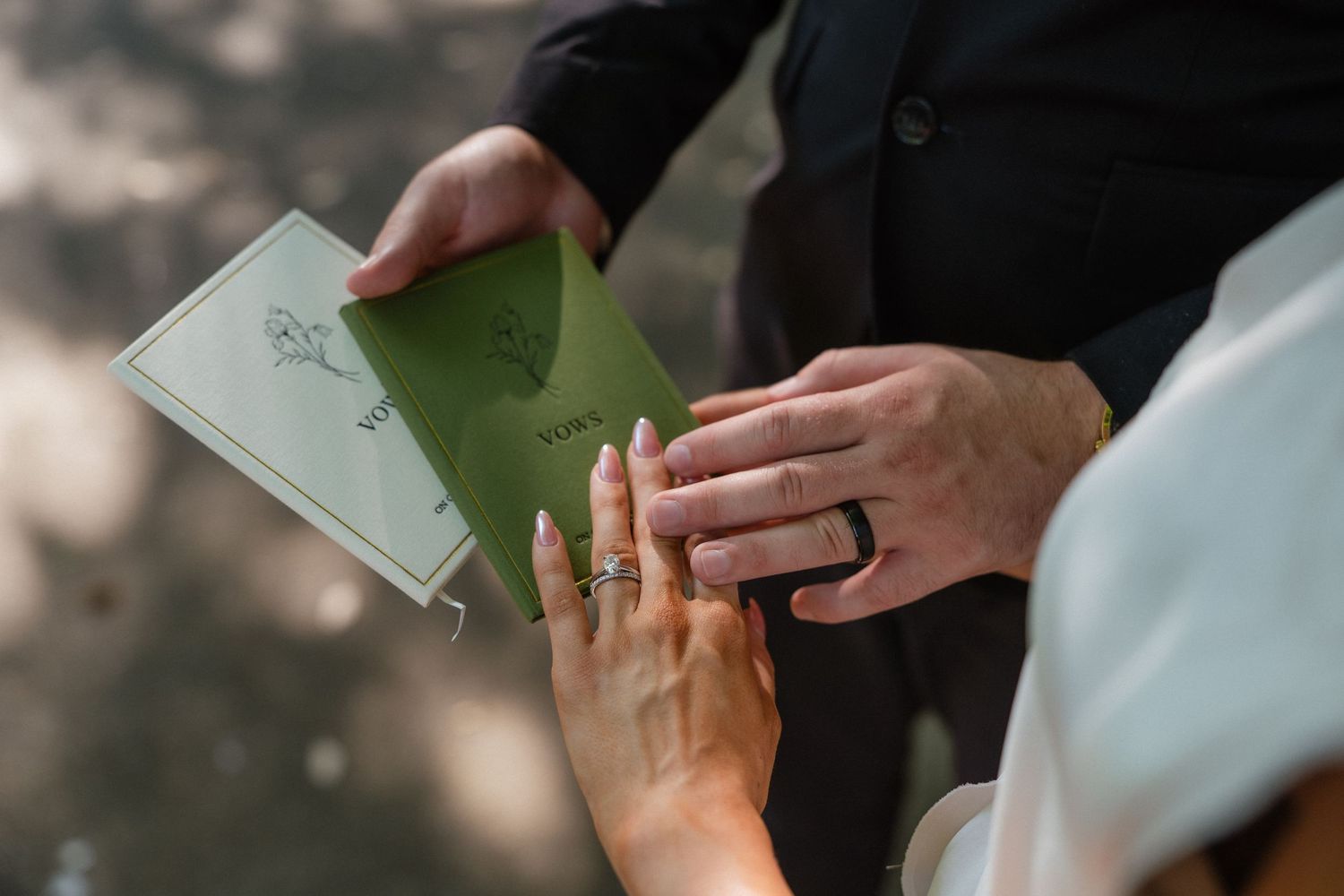 Close-up of couple exchanging wedding rings while groom holds green vows booklet during outdoor ceremony.