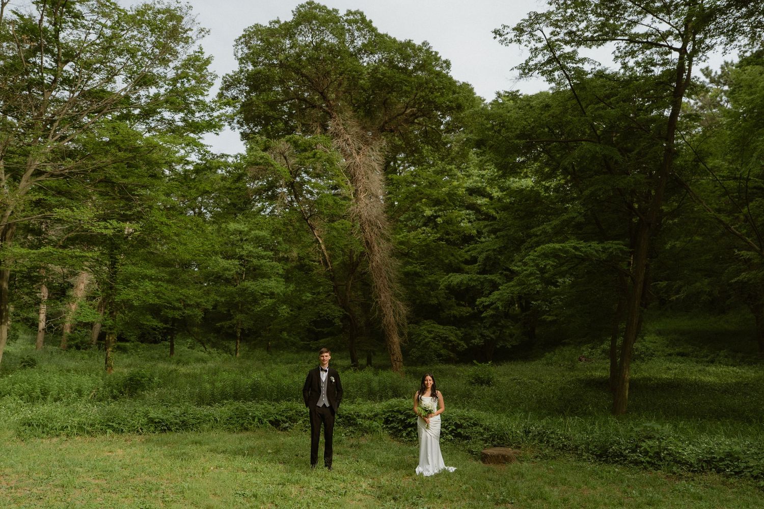 Bride and groom stand apart in a lush green forest clearing during an outdoor wedding ceremony.
