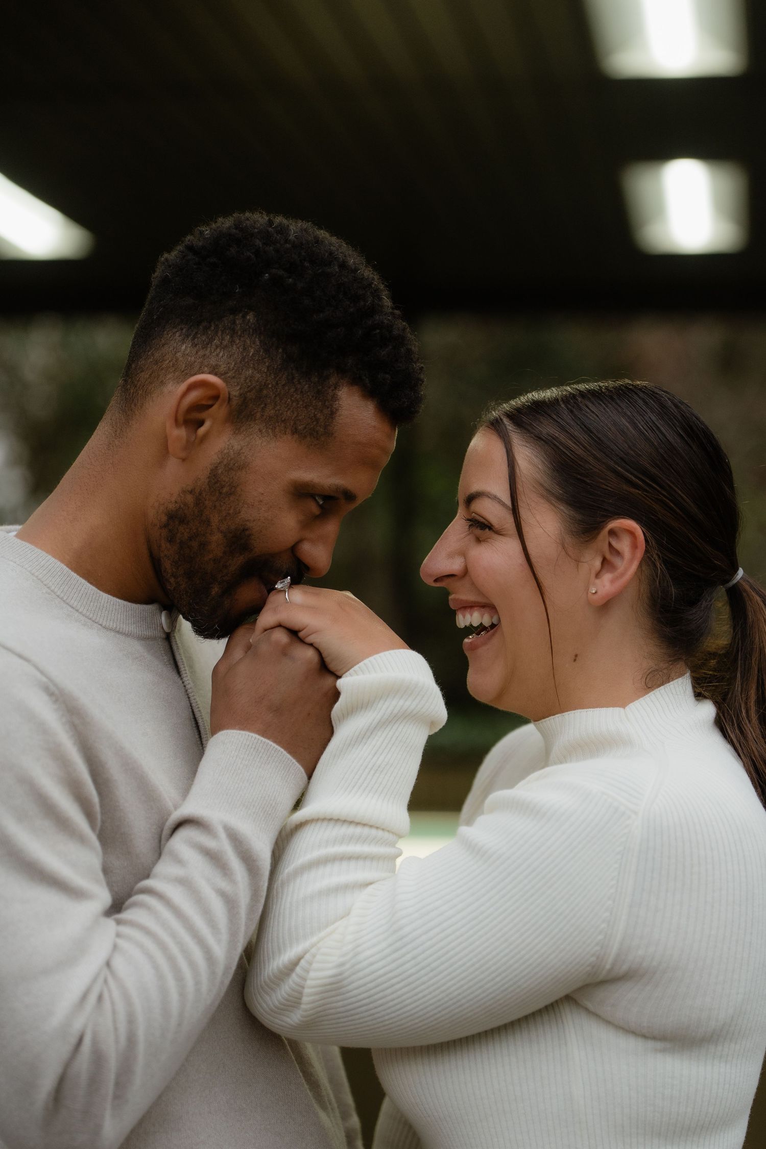 A joyful interracial couple in matching white sweaters share a tender moment, the man kissing the woman's hand as she laughs.