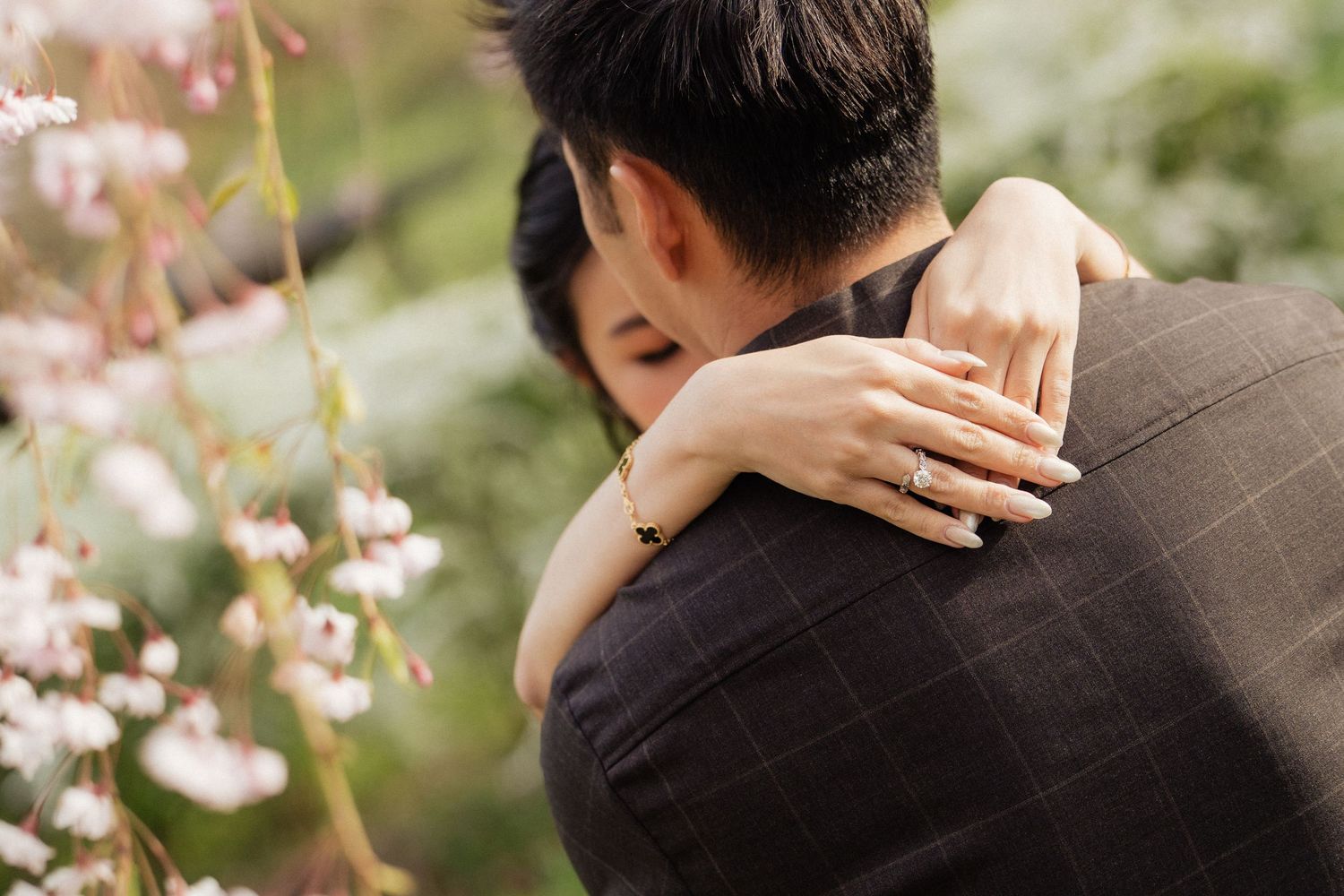 Couple embracing among cherry blossoms, woman showing diamond engagement ring over man's shoulder.