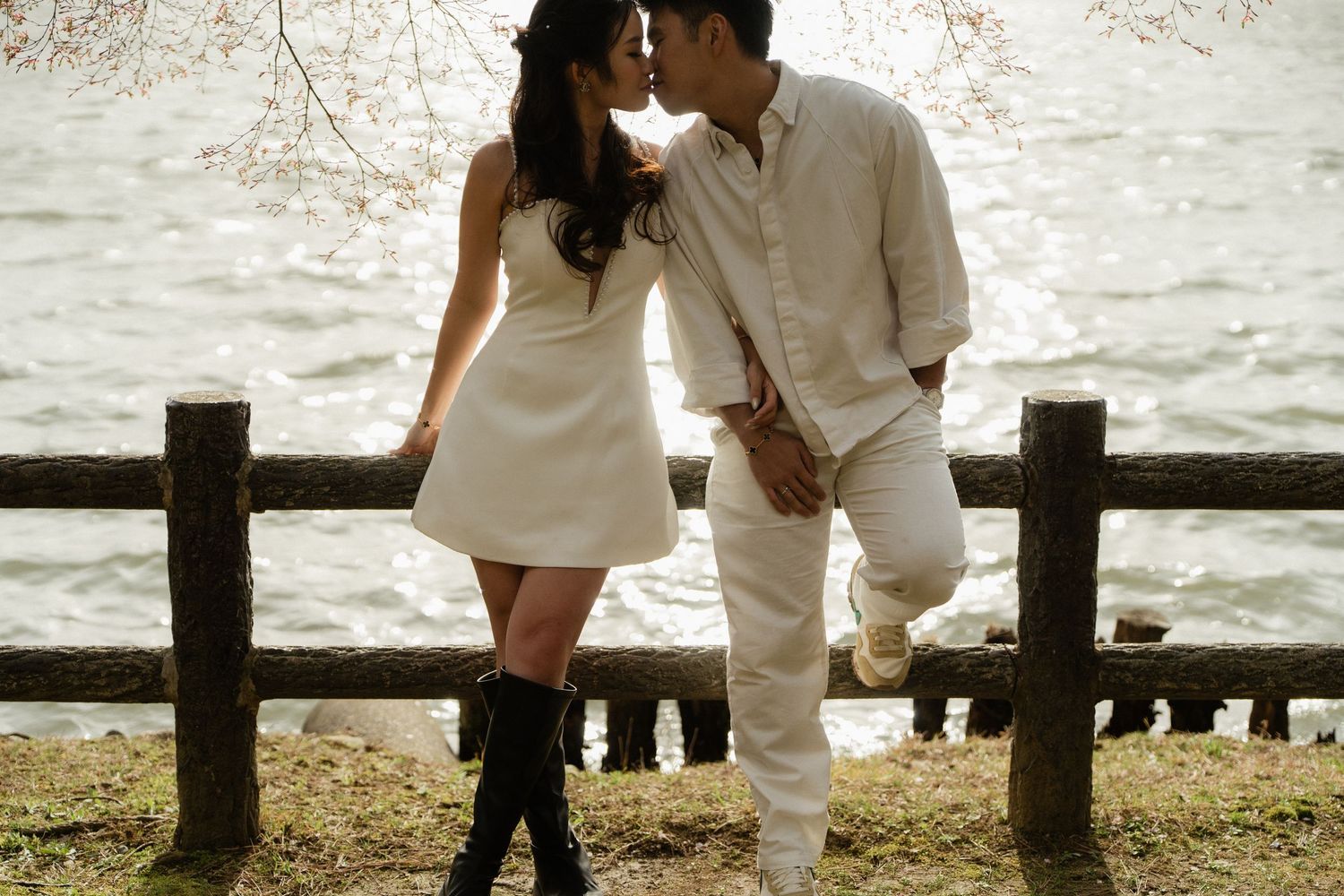 Couple sharing a romantic moment by a lakeside fence surrounded by cherry blossom trees in spring.