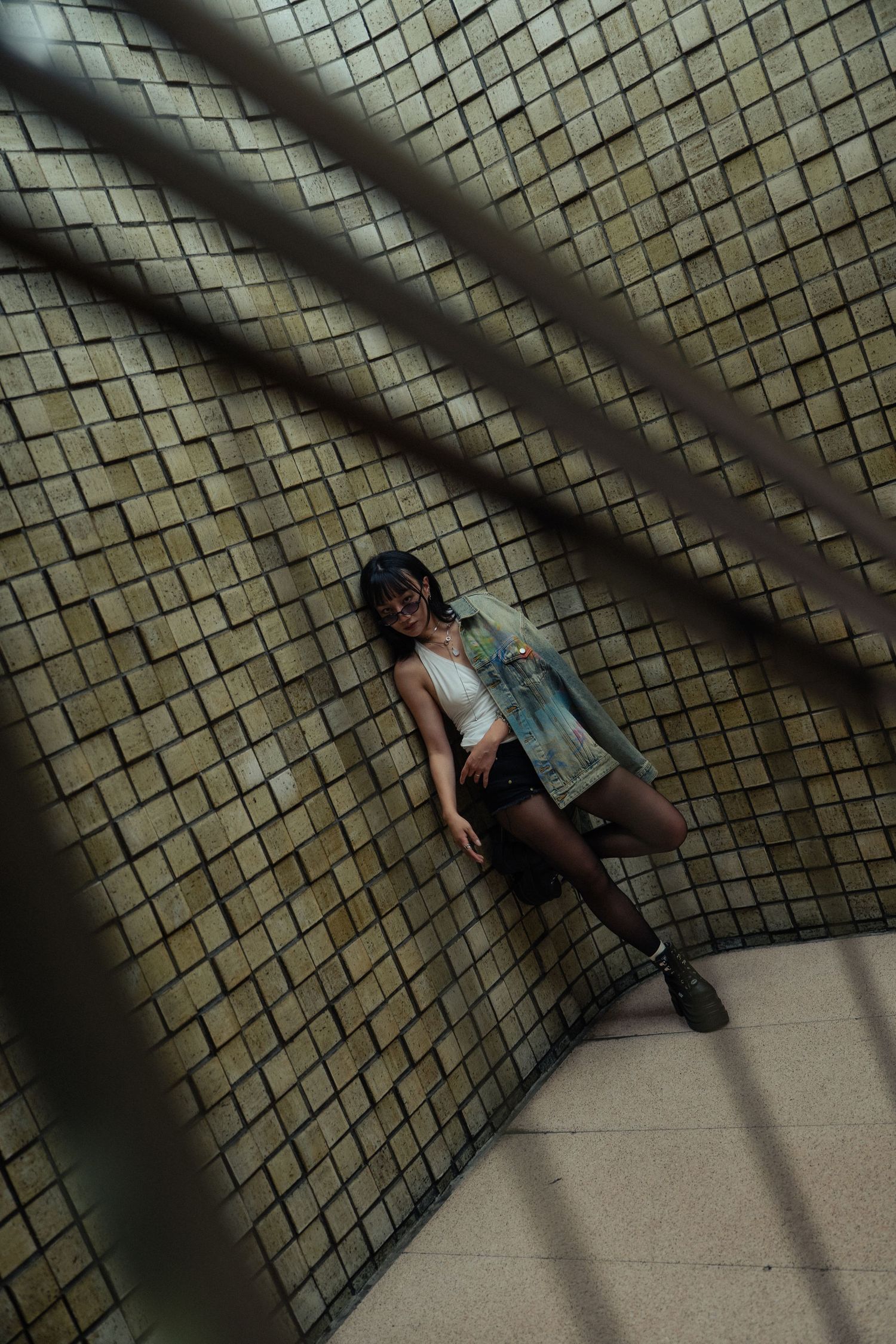 Young woman in denim jacket and mini skirt posing against tiled wall in urban underground setting.