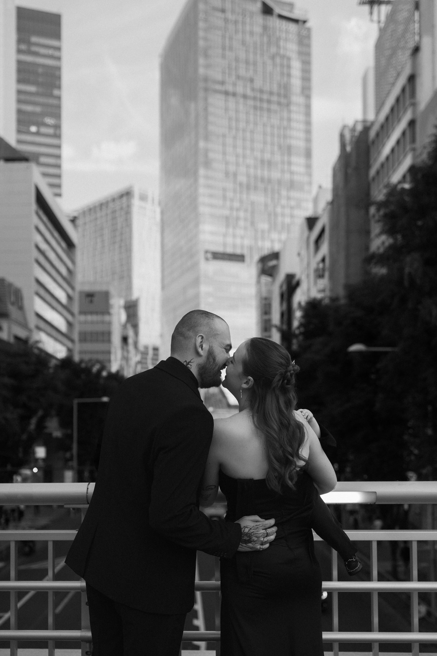 Black and white photo of a couple kissing on a bridge with city skyscrapers in the background.