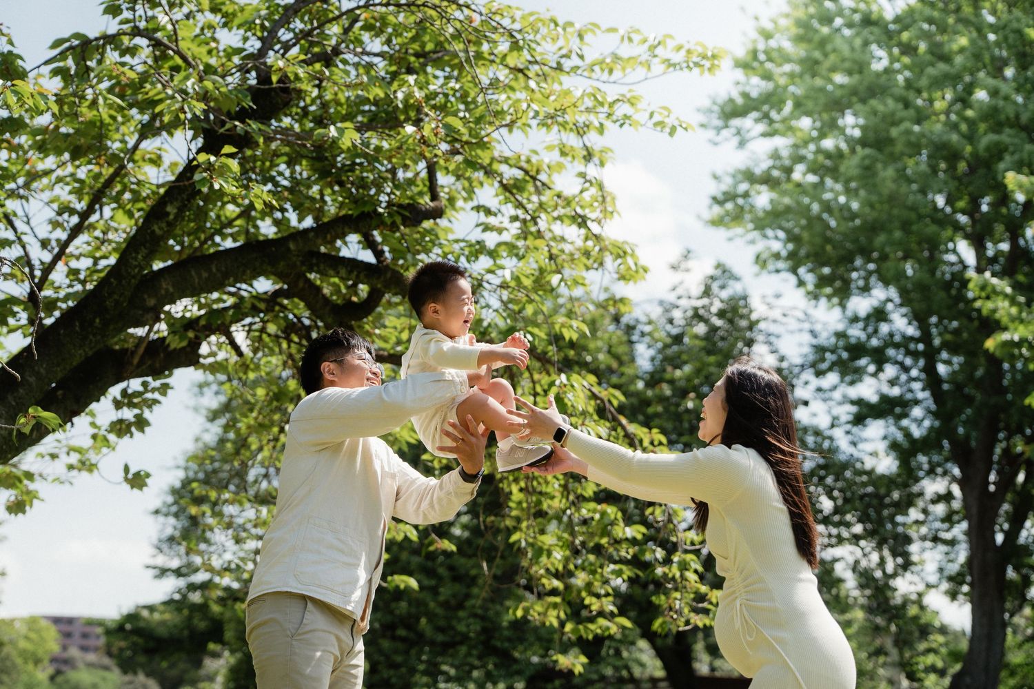 Happy Asian family playing outdoors, parents lifting joyful child in sunny green park.